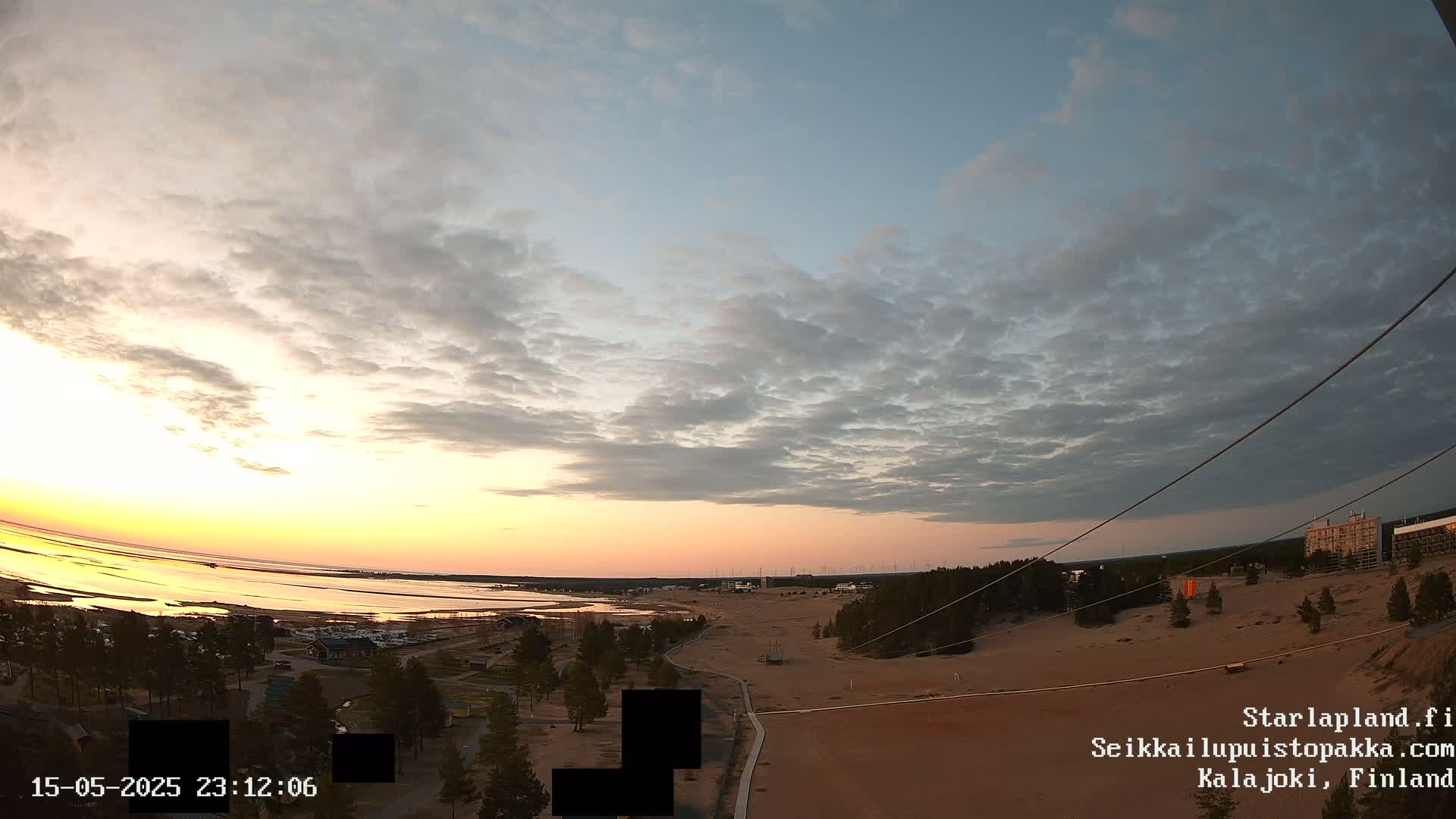 A wide, high-angle view shows a sunset over a calm body of water, with a sandy area and trees in the foreground under a partly cloudy sky.
