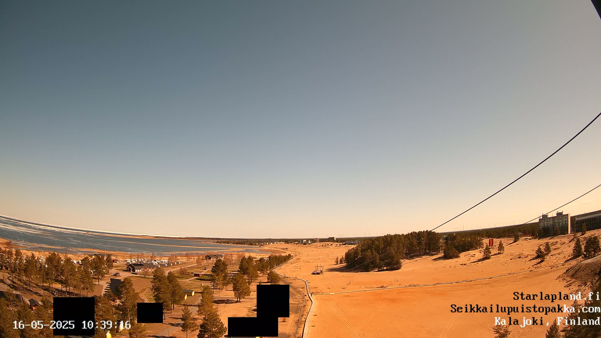 A mostly clear, bright day reveals a sandy landscape with sparse trees, a body of water with ice along the shore, and a paved path leading to distant buildings.