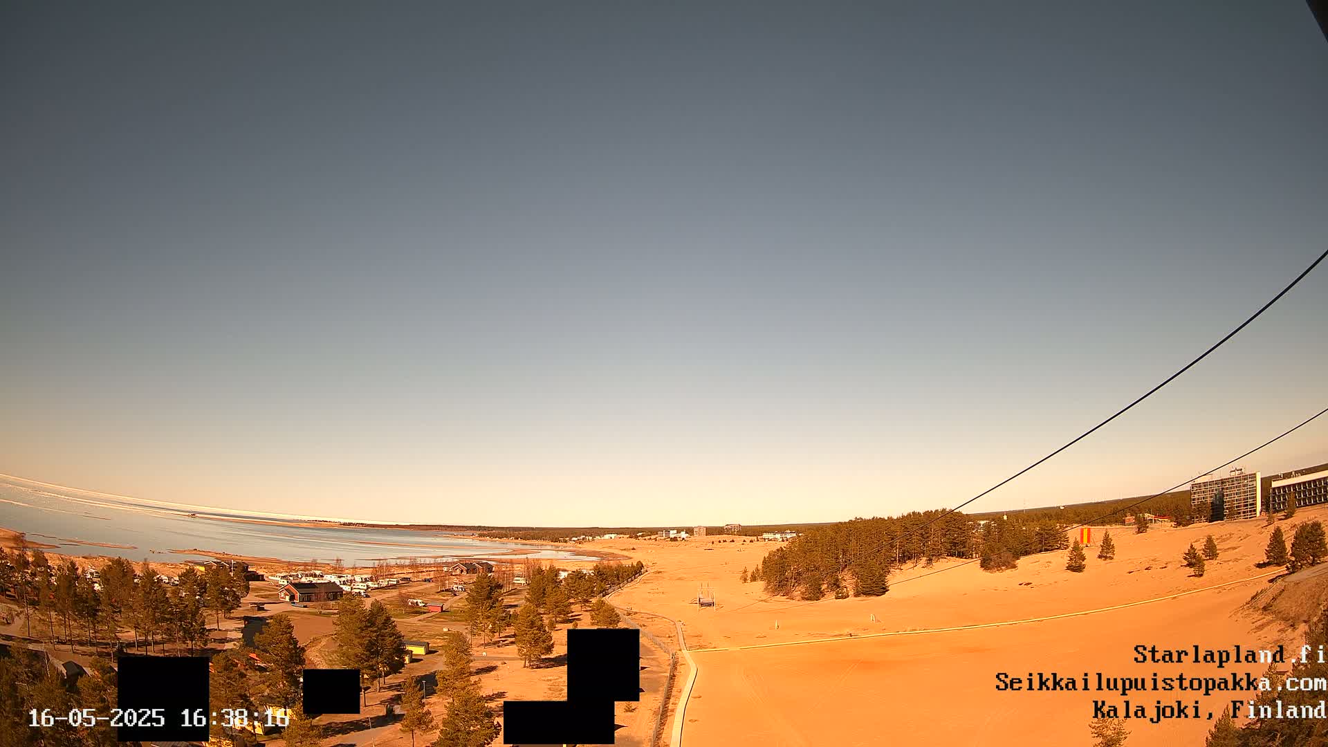 A mostly clear sky overlooks a sandy landscape with sparse vegetation, a body of water partially covered in ice, and buildings in the distance.