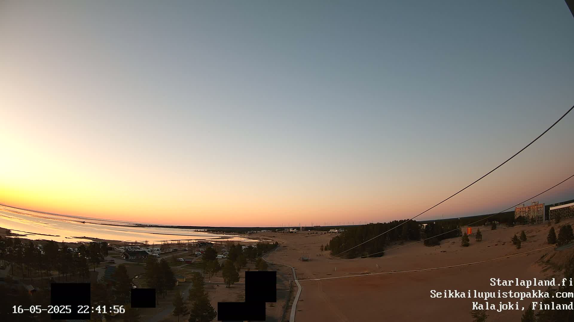 A wide shot at sunset shows a sandy area with sparse vegetation near a calm body of water, under a clear sky.