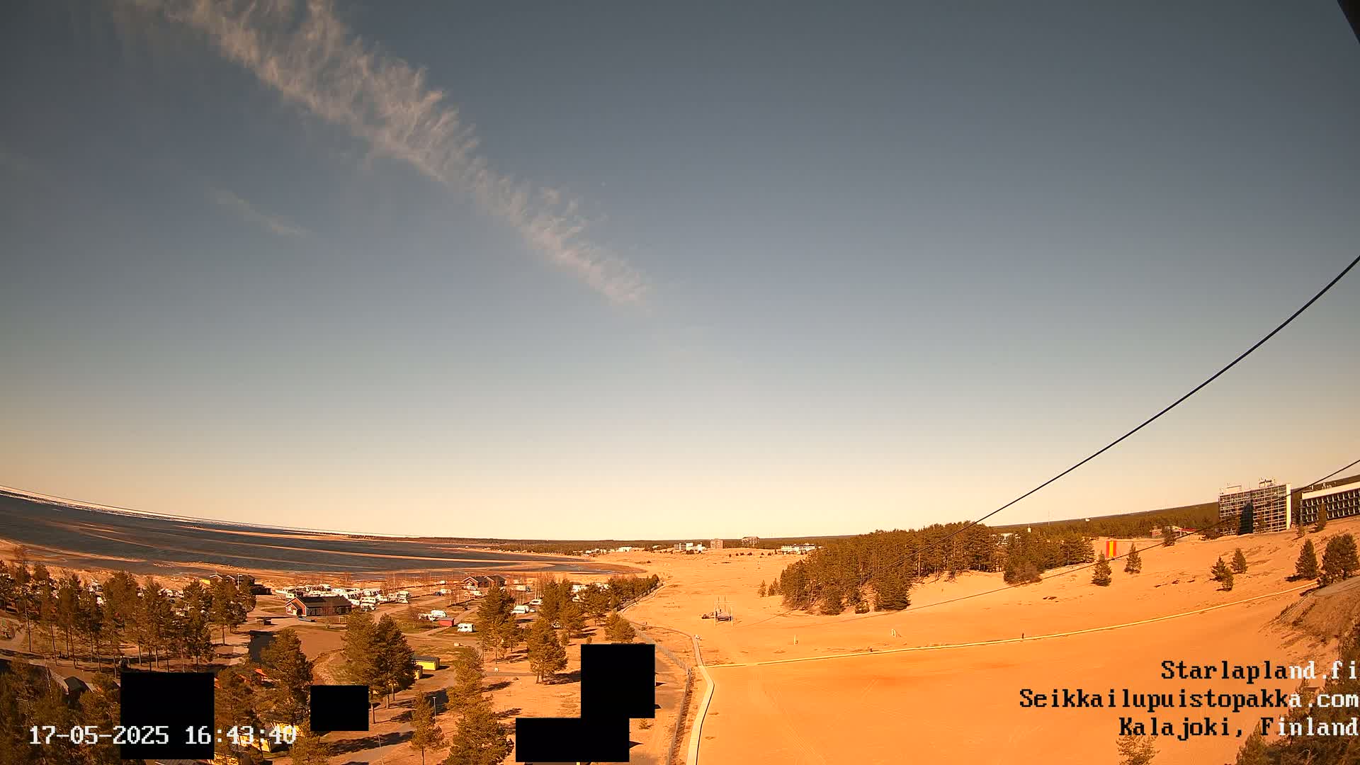 A high-angle view shows a sandy landscape with sparse vegetation, a body of water in the distance, and a mostly clear sky with a thin, wispy cloud formation.