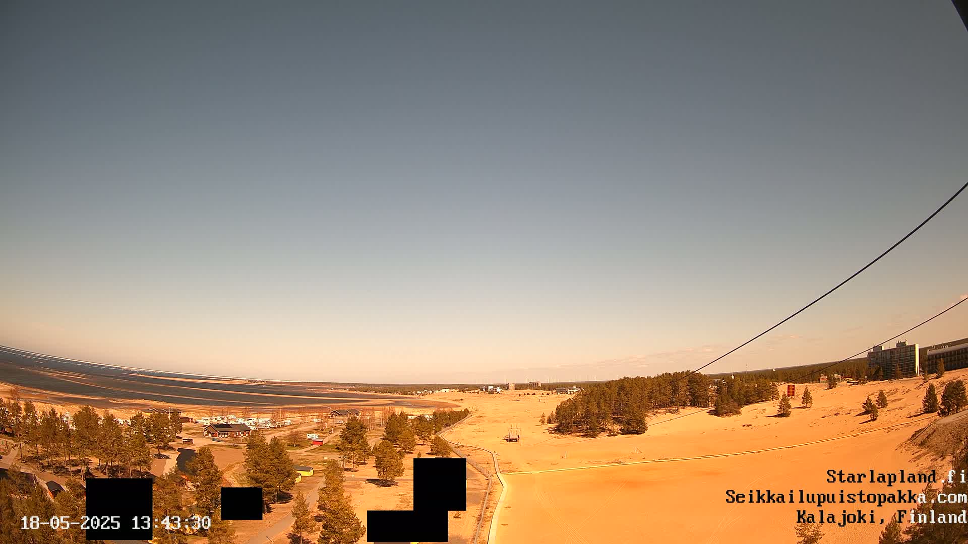 A high-angle view shows a sandy landscape with sparse vegetation, a body of water in the distance, and a clear, light-colored sky.
