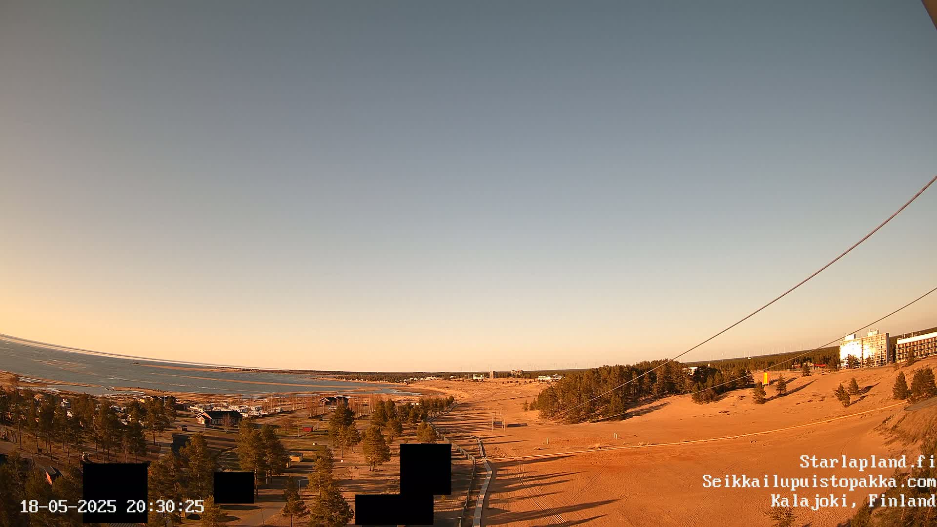 A high-angle, long shot shows a sandy beach and calm body of water at sunset, with a line of trees separating the beach from a residential area.