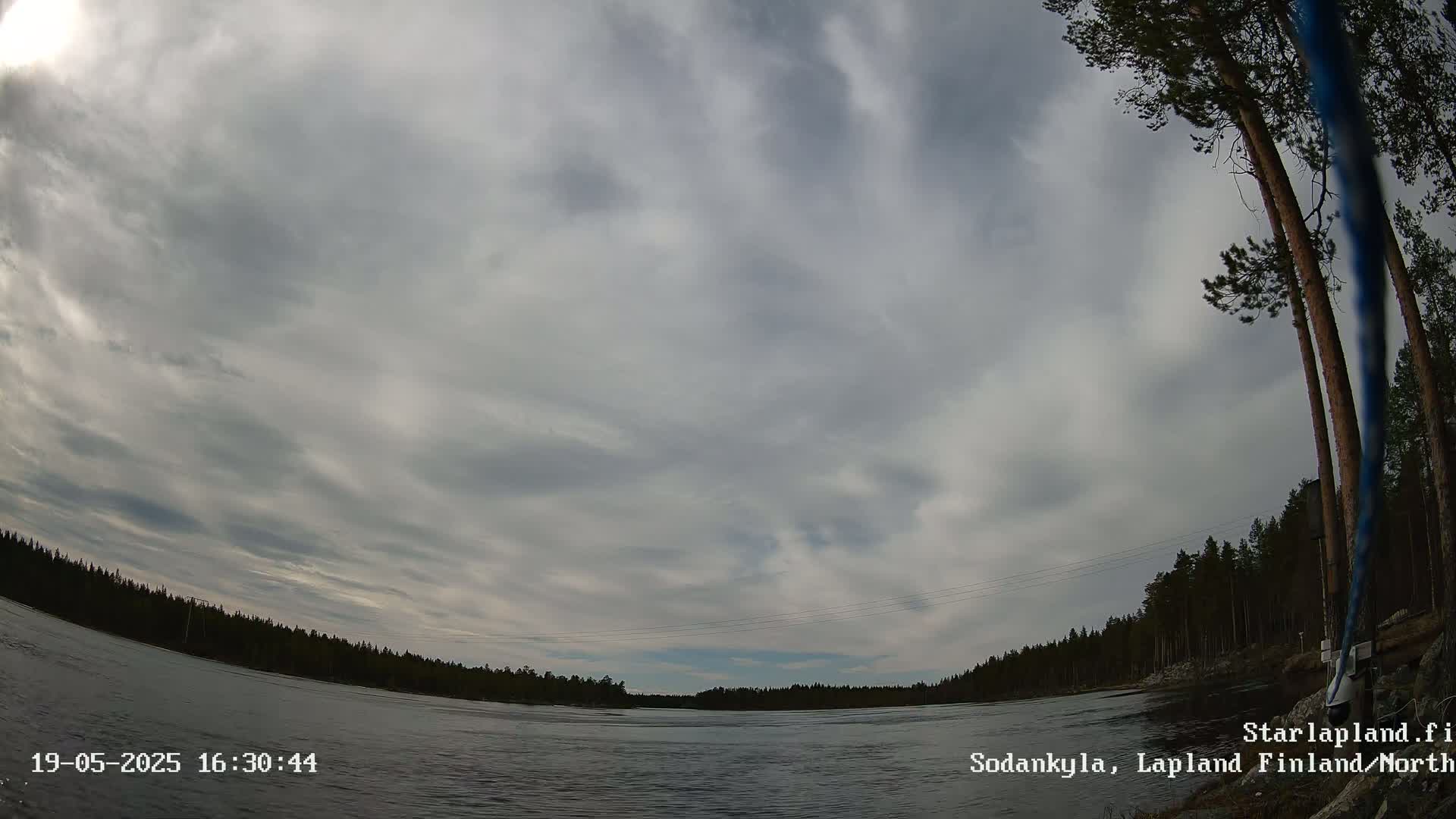 A wide shot shows a mostly cloudy sky over a calm river, bordered by a dark forest under a light overcast.