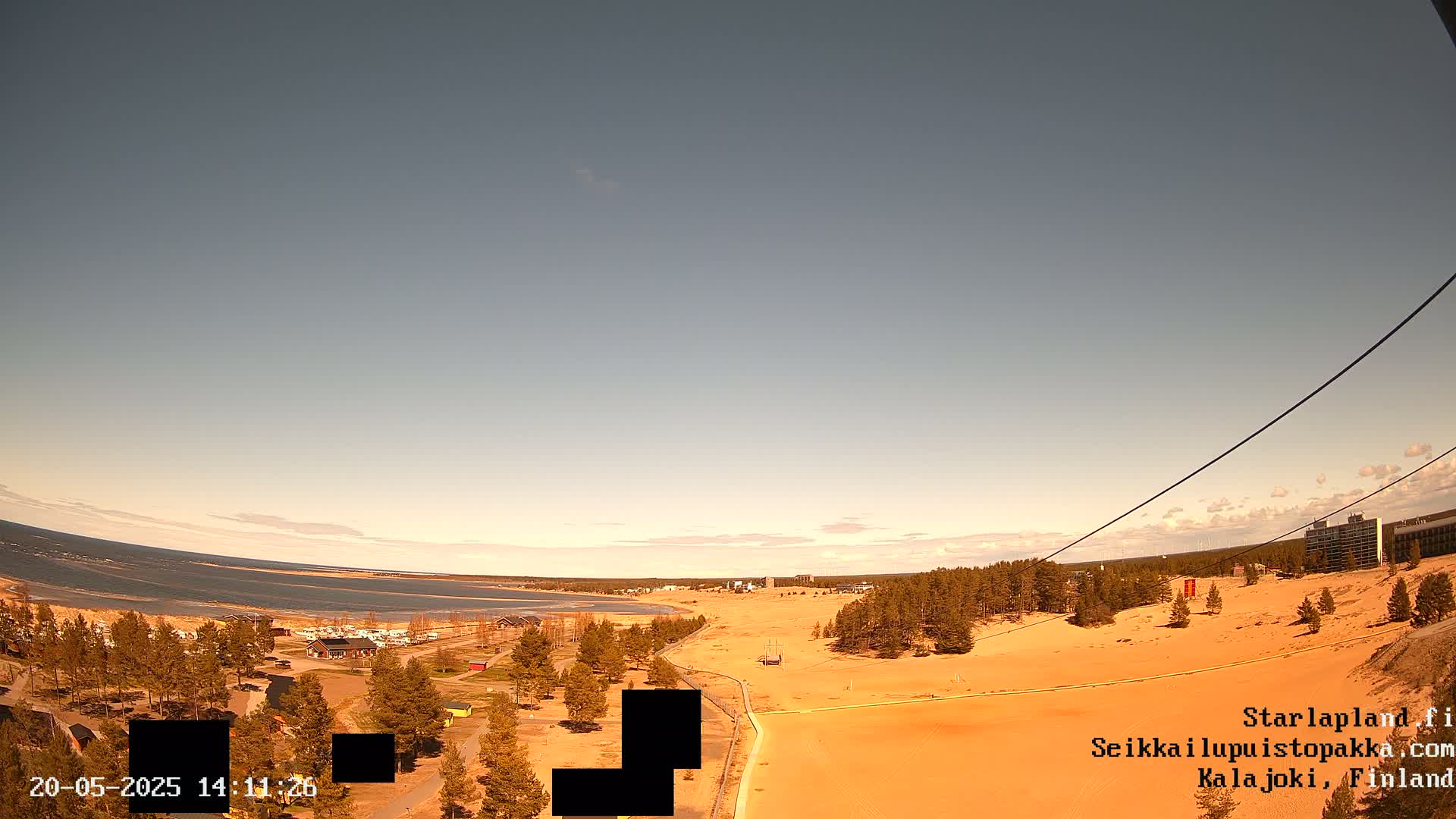 A high-angle, long shot shows a sandy landscape next to a calm body of water under a mostly clear sky.
