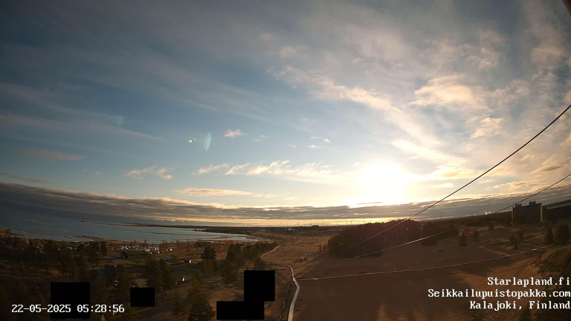 A partly cloudy sunset over a coastal landscape with a sandy area, some trees, and buildings in the distance.