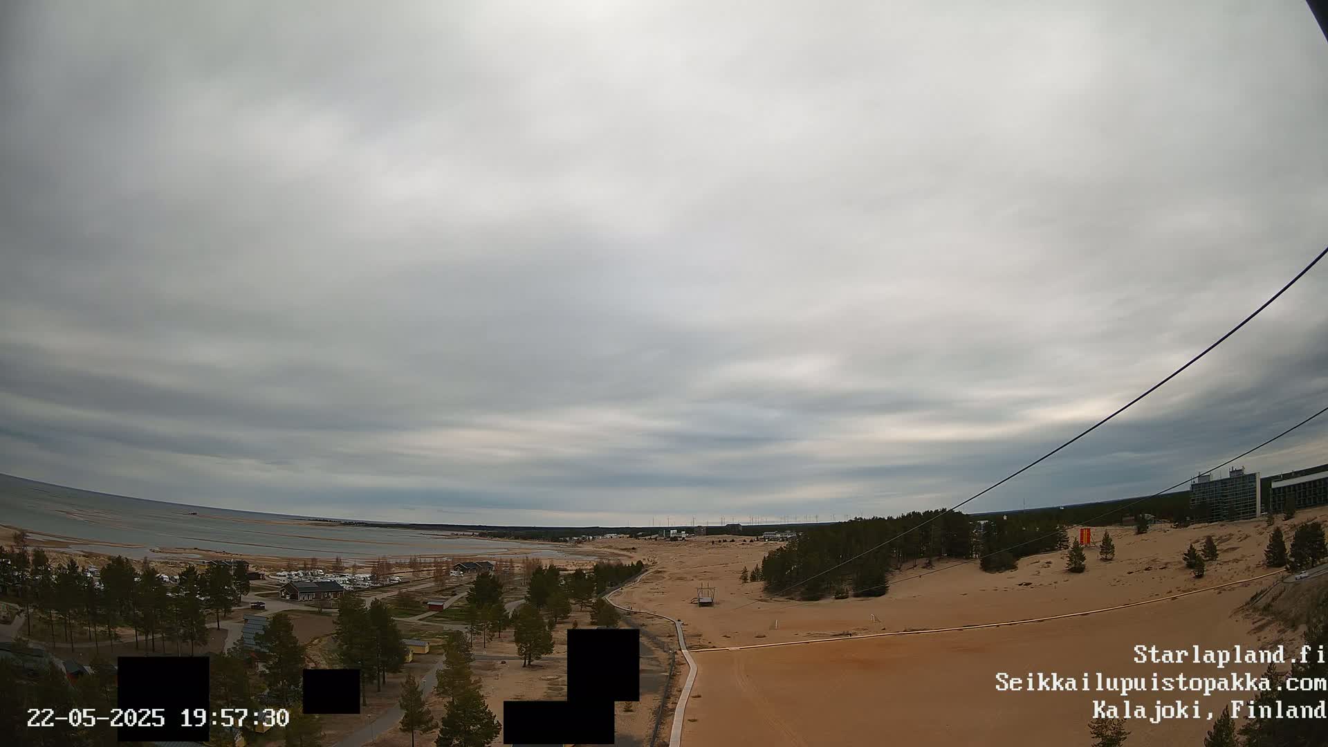 An overcast sky overlooks a sandy landscape with a body of water in the distance, trees, and buildings.