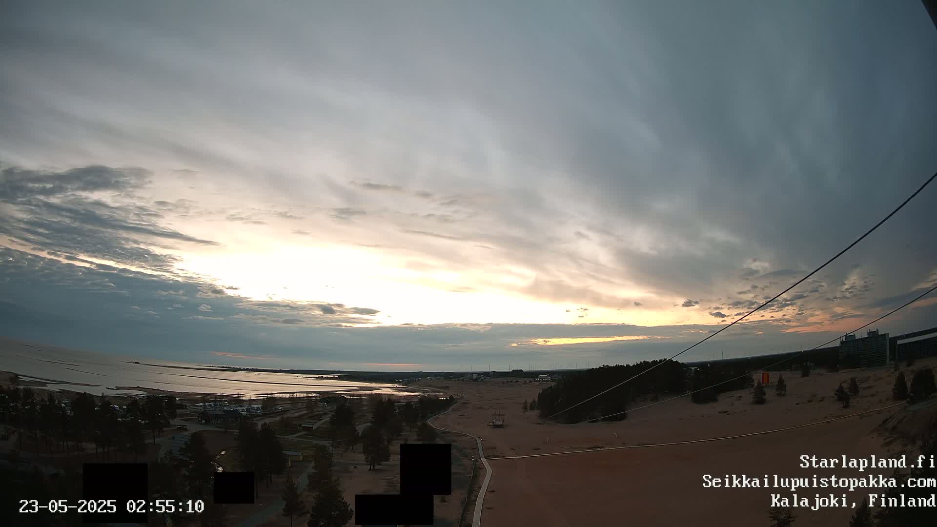 A wide, high-angle view shows a calm body of water under a cloudy sunset sky over a sandy landscape with sparse vegetation and a few buildings in the distance.