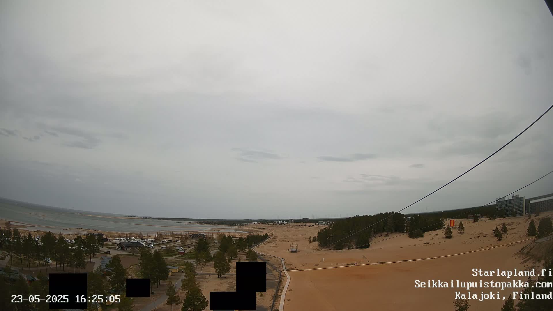An overcast sky hangs over a sandy landscape with a body of water in the distance, featuring sparse vegetation, buildings, and a zipline cable.