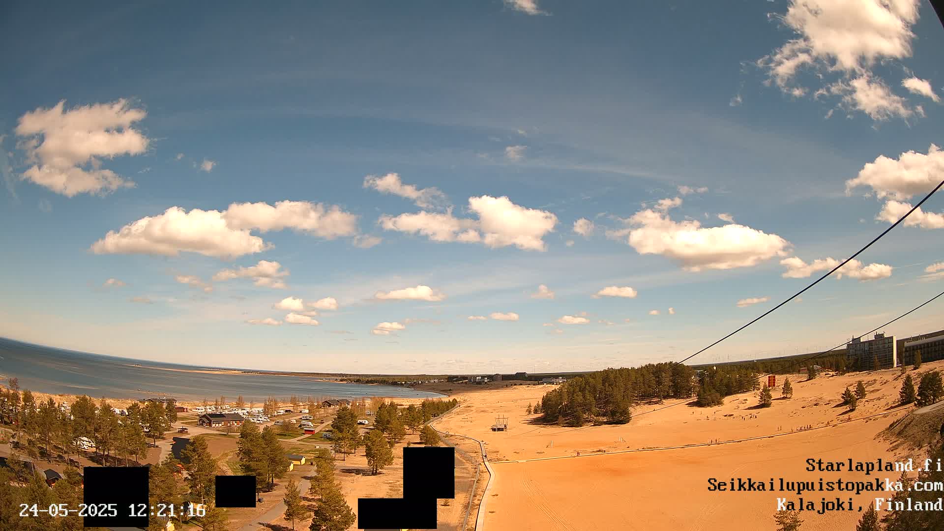 A high-angle view shows a sandy area next to a calm body of water under a partly cloudy sky.