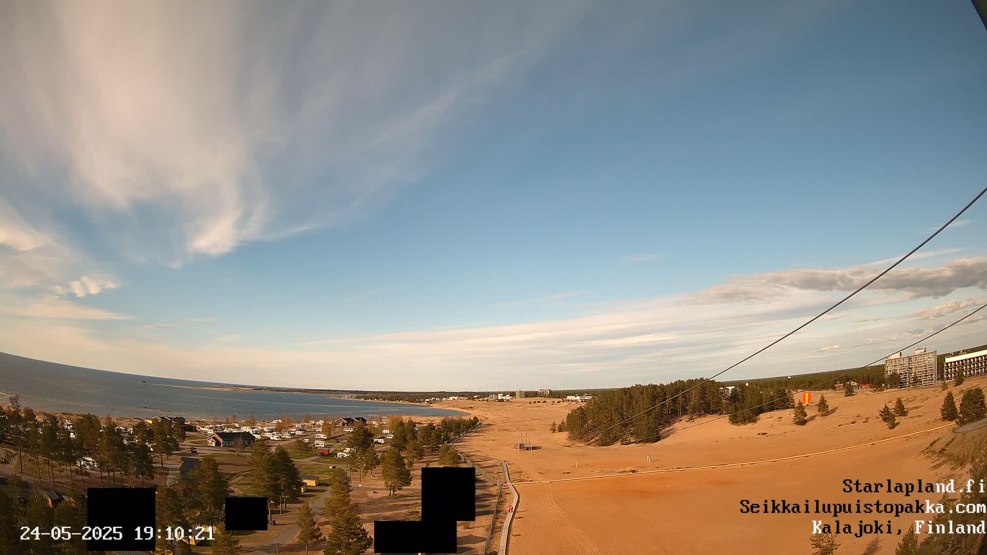 A mostly sunny day reveals a sandy shoreline with a calm body of water in the background, sparsely populated with buildings and trees.