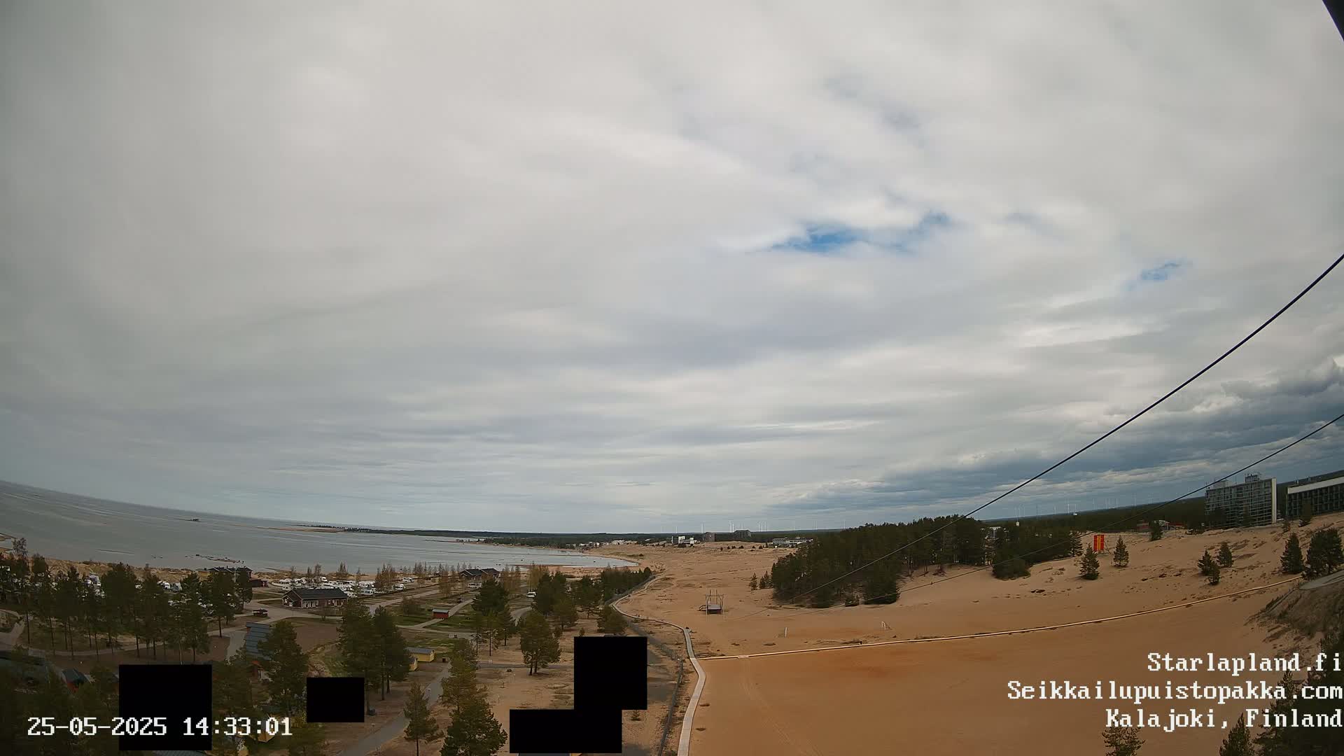 A mostly cloudy day overlooks a sandy beach and calm sea with sparse vegetation and buildings visible in the distance.