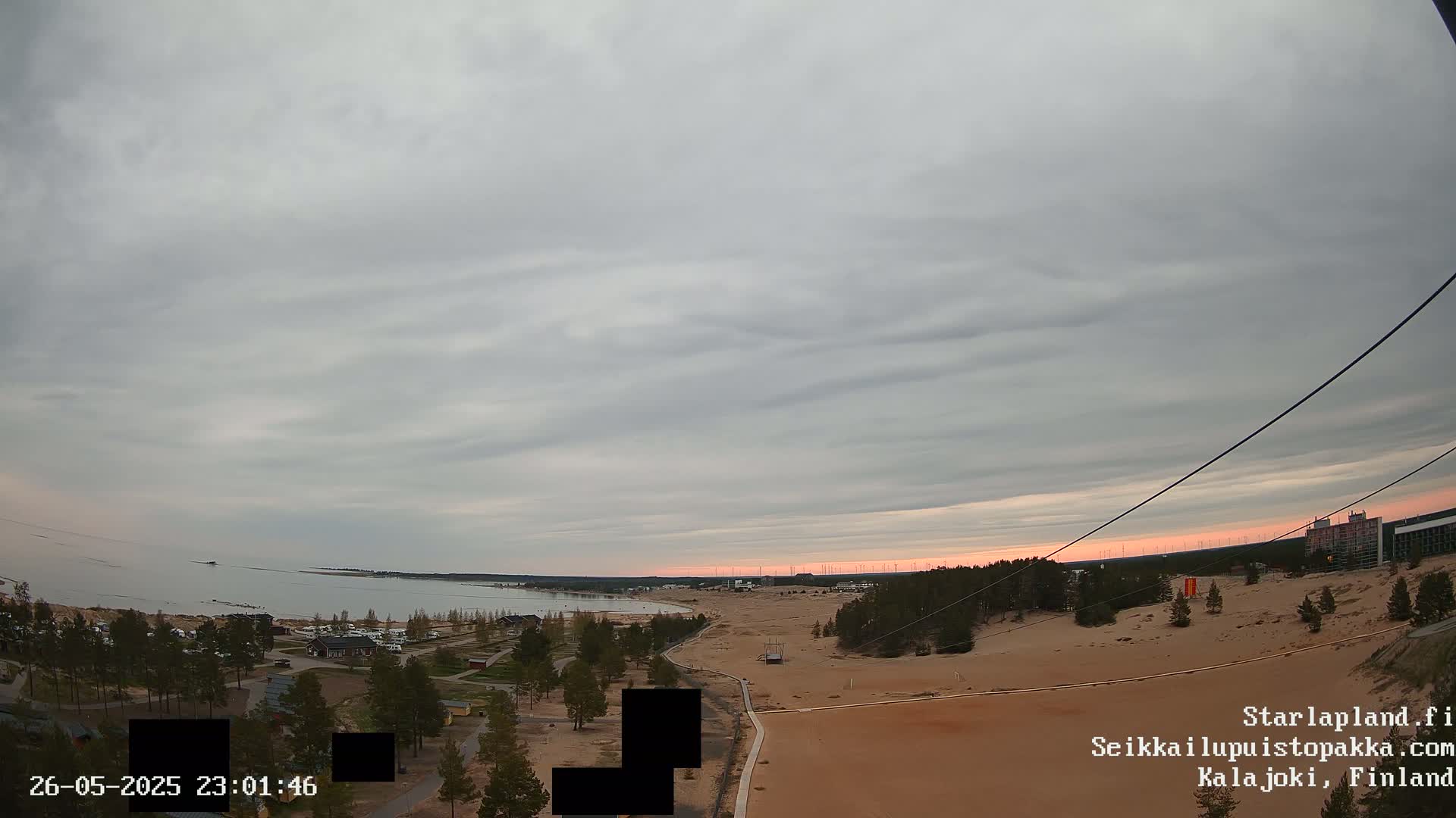 An overcast sky with a hint of pink at the horizon overlooks a sandy landscape, a calm body of water, and a sparse collection of buildings.