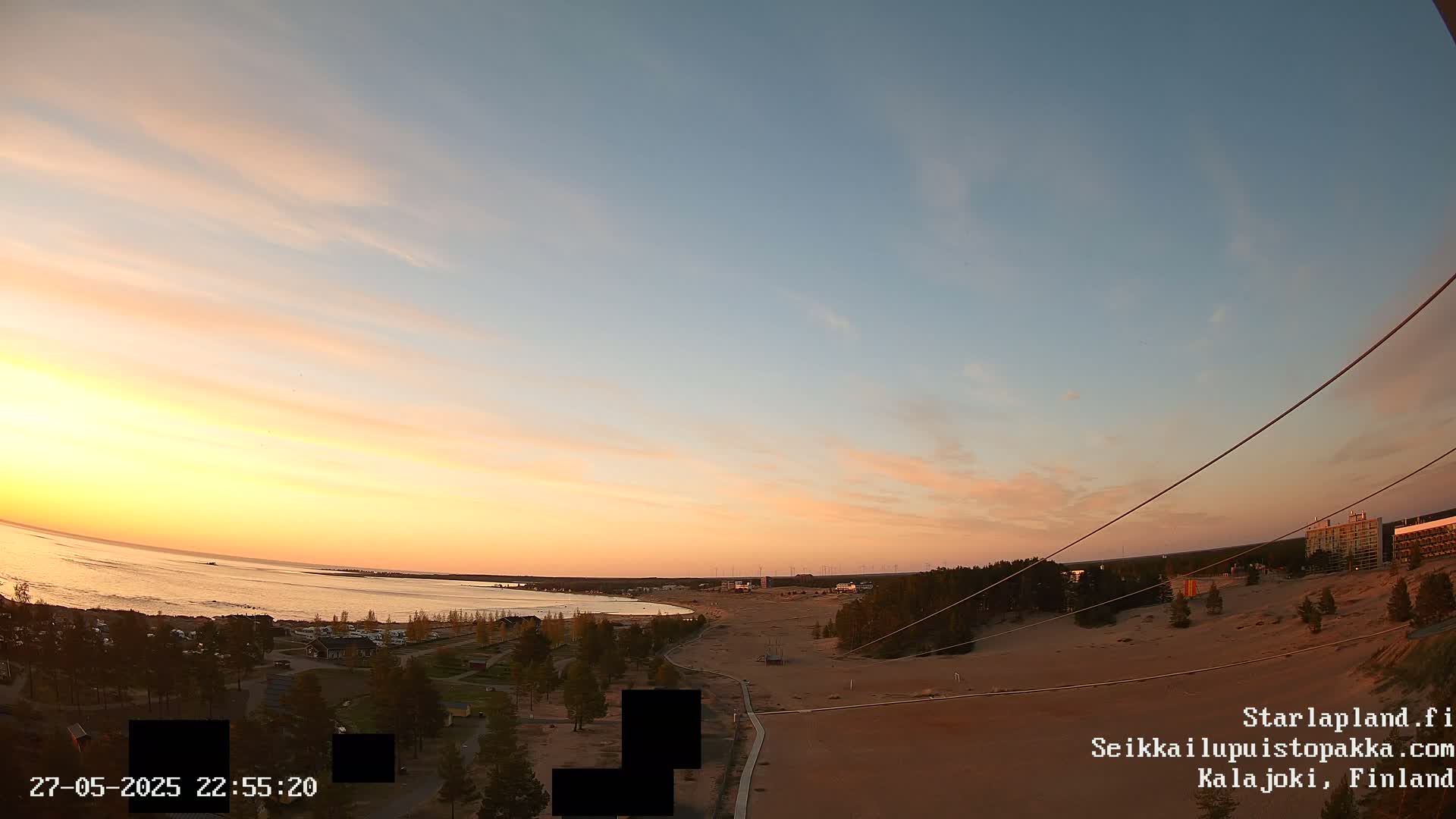 A sunset over a sandy beach and calm sea, with sparse trees and buildings in the distance.
