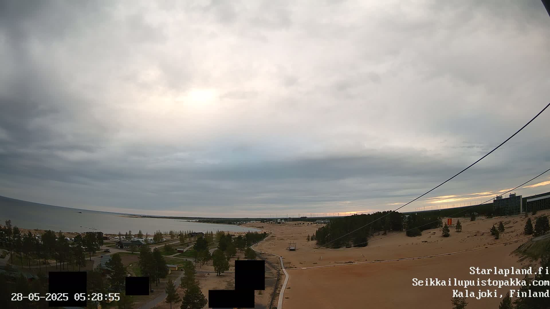 An overcast sky hangs over a sandy landscape with a calm body of water in the background, and some sparse vegetation and buildings are visible.