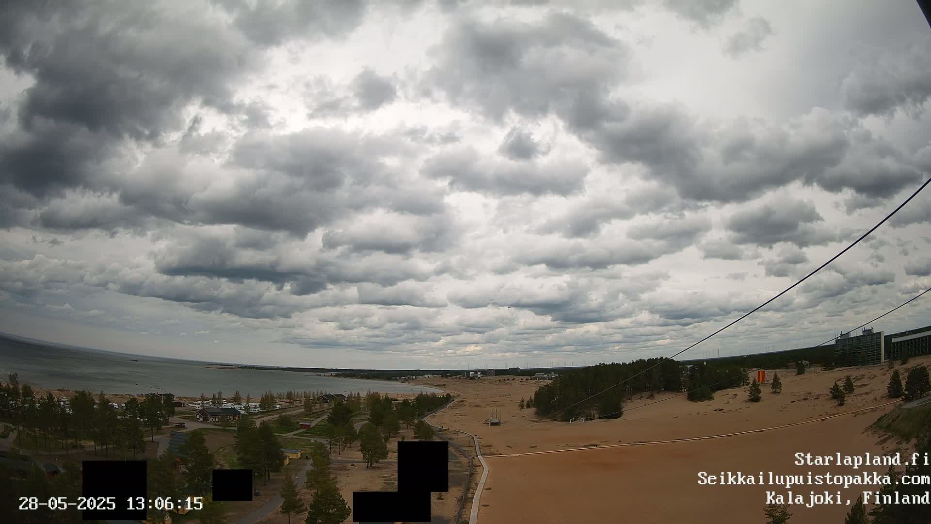 A mostly cloudy sky overlooks a sandy landscape next to a body of water with some trees and buildings in the distance.