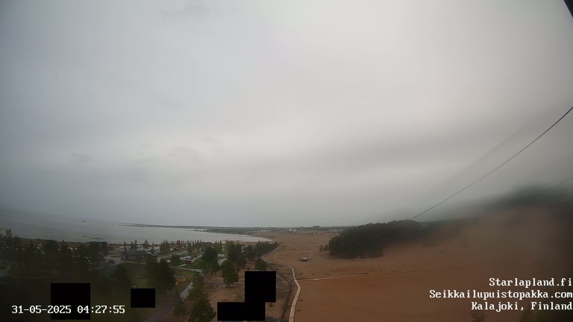 A high-angle, overcast view shows a calm body of water, a sandy shore, and sparse vegetation under a gray sky.