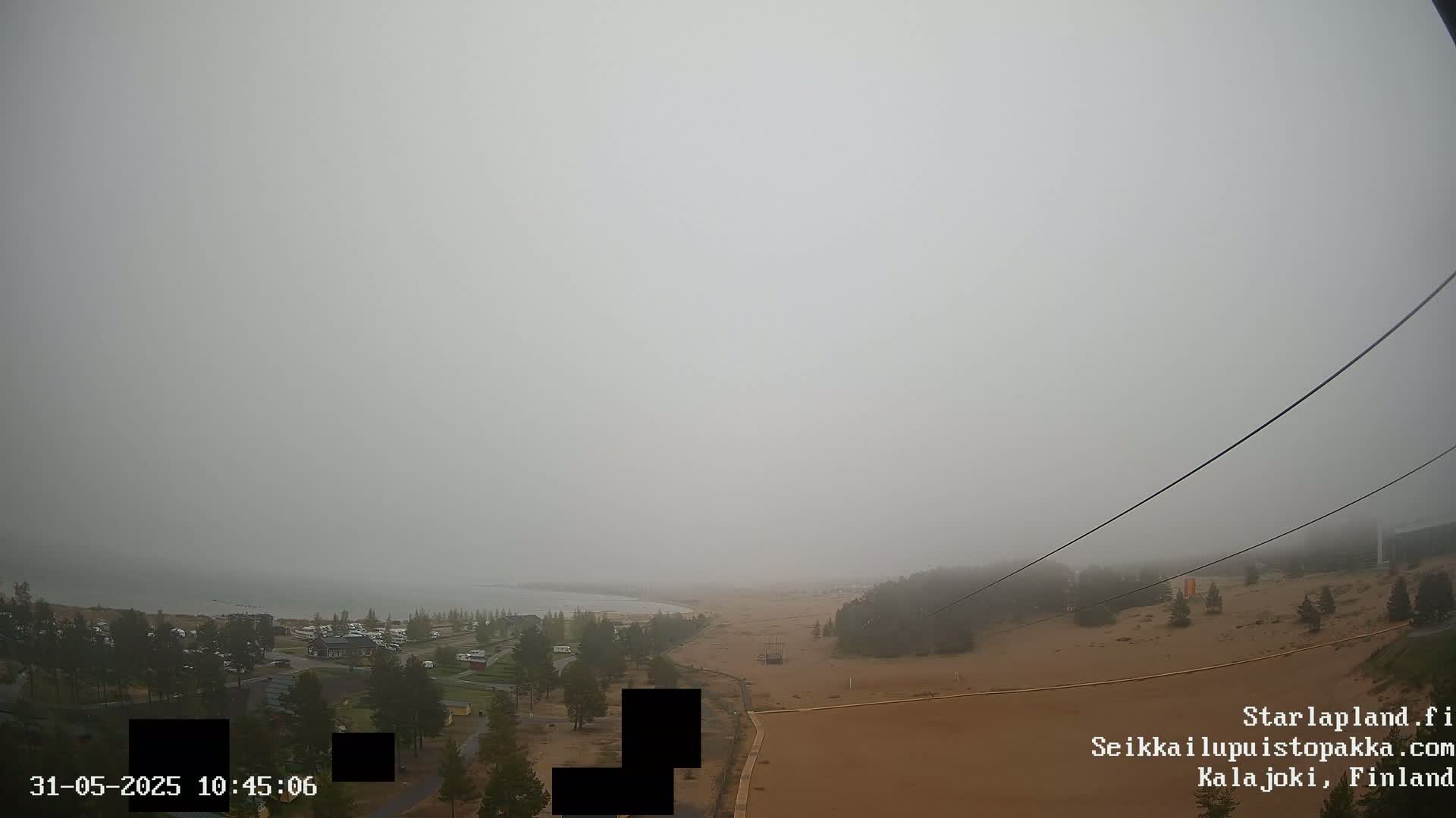 A foggy coastal scene shows a sandy area with sparse vegetation and a body of water in the distance, viewed from above.
