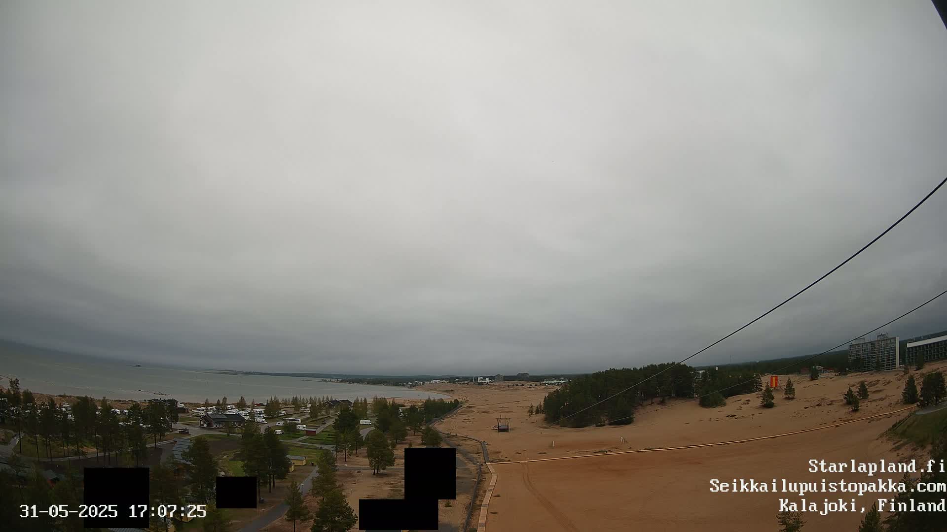 A high-angle view shows a grey, overcast day over a sandy landscape next to a calm body of water, with trees, buildings, and a zipline visible in the distance.