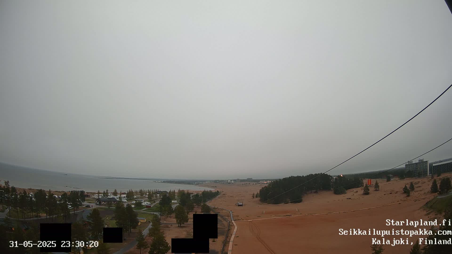 An overcast sky hangs over a sandy landscape featuring a body of water, trees, buildings, and what appears to be a zip line.