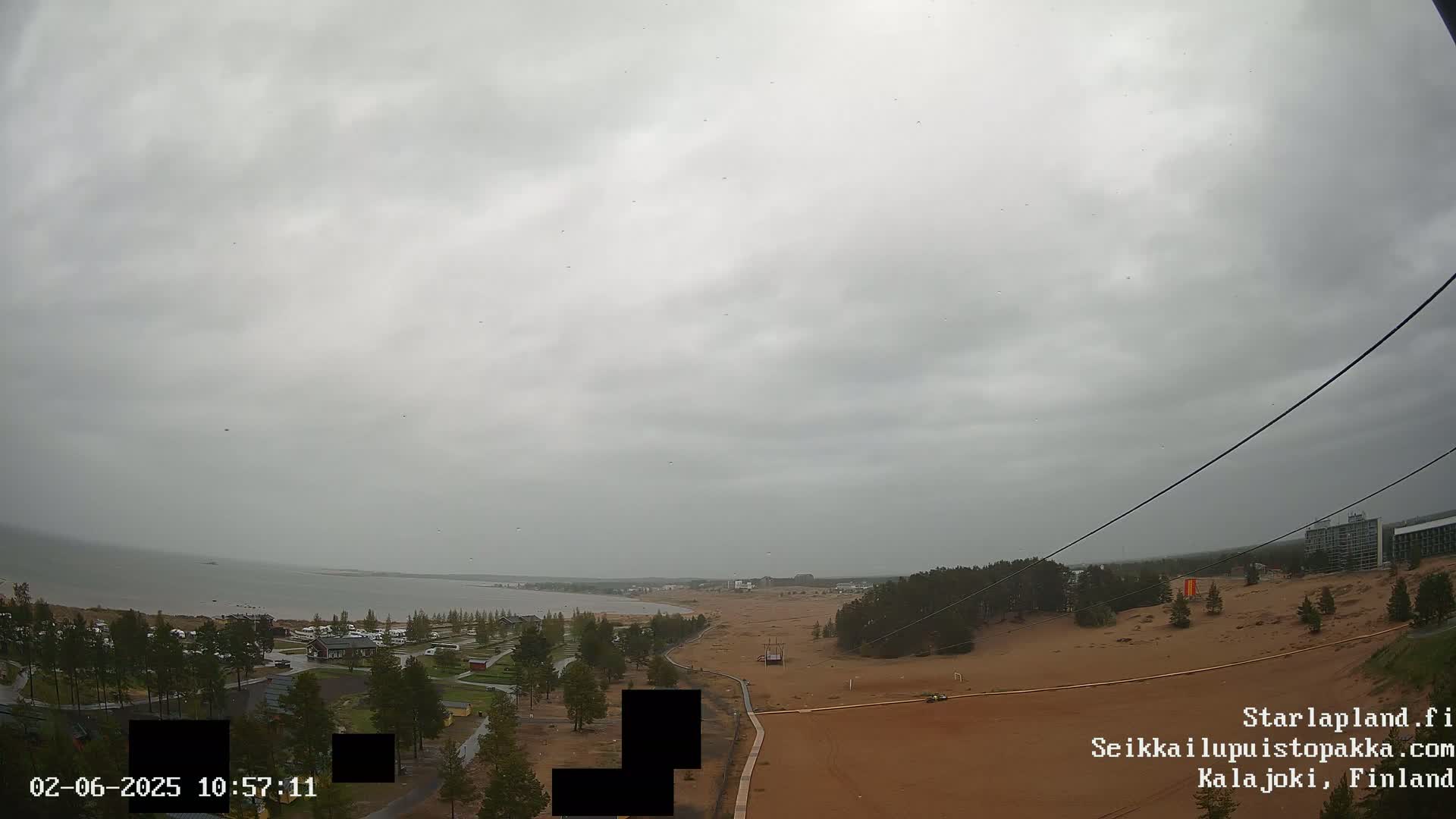 An overcast sky over a sandy landscape with a body of water in the distance,  a few buildings, and some vegetation.