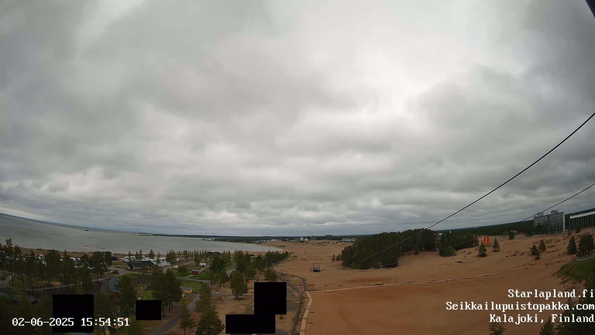 An overcast sky hangs over a sandy landscape next to a calm body of water, with sparse trees and buildings visible in the distance.