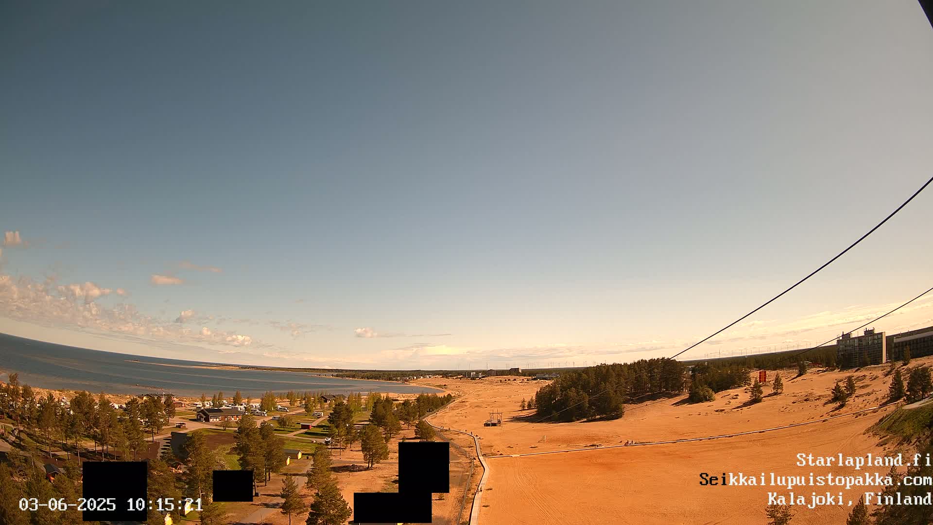 A mostly clear sky overlooks a sandy landscape with a body of water and sparse vegetation, including a small campground, under bright, sunny conditions.