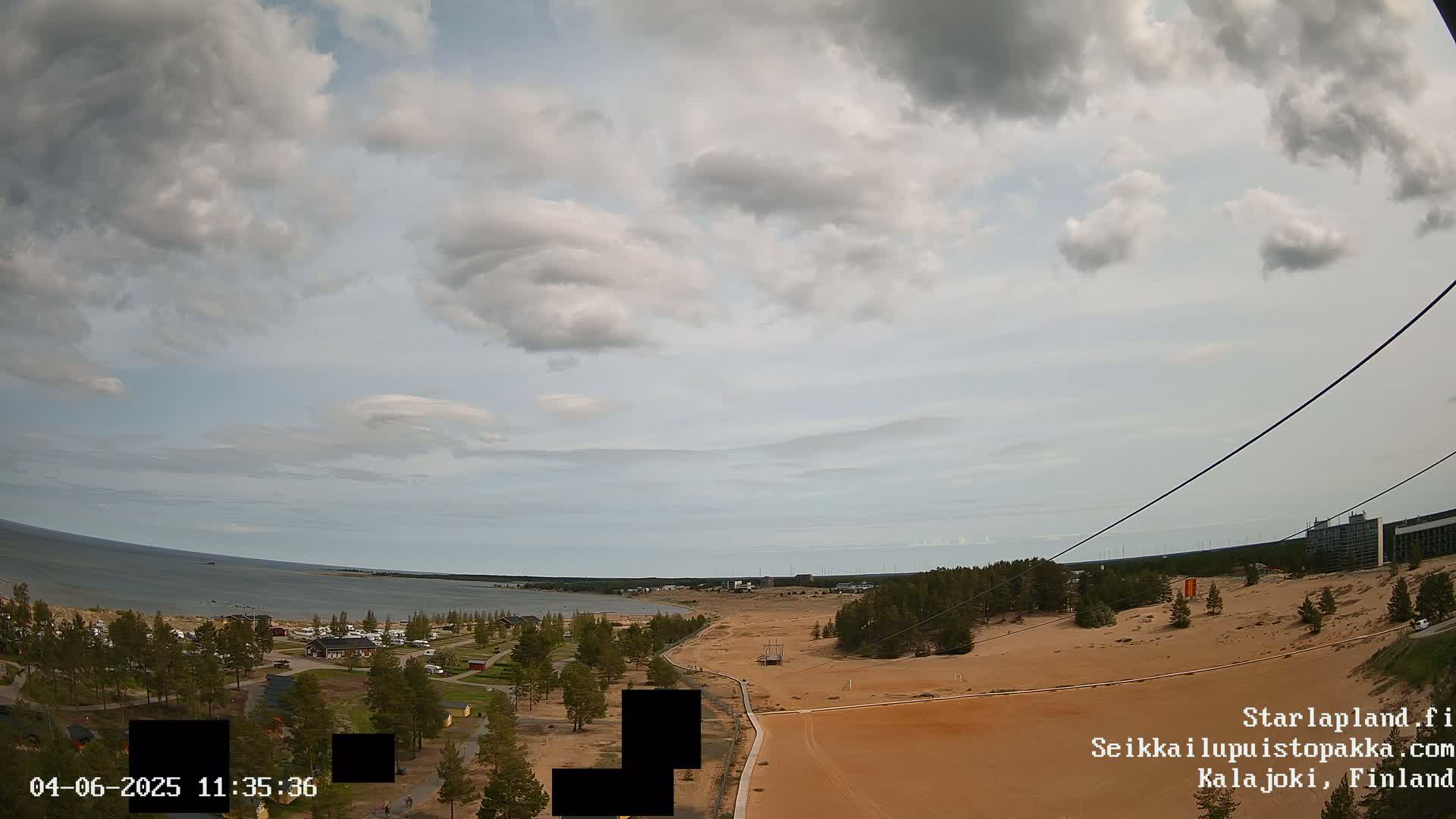 A mostly cloudy sky overlooks a sandy landscape with a body of water and some buildings in the distance, viewed from a high vantage point with cables visible.