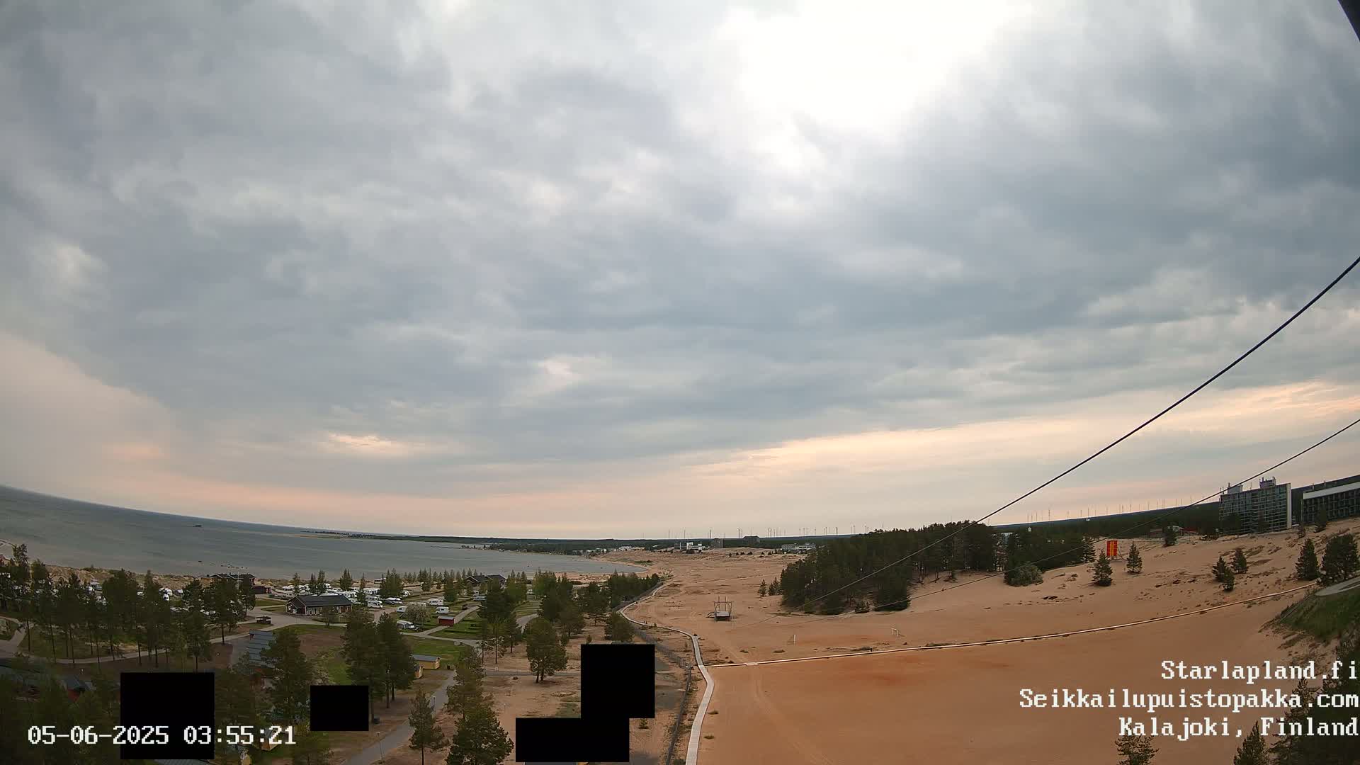 An overcast sky hangs over a sandy landscape with a body of water in the distance, featuring some trees, buildings and a path.