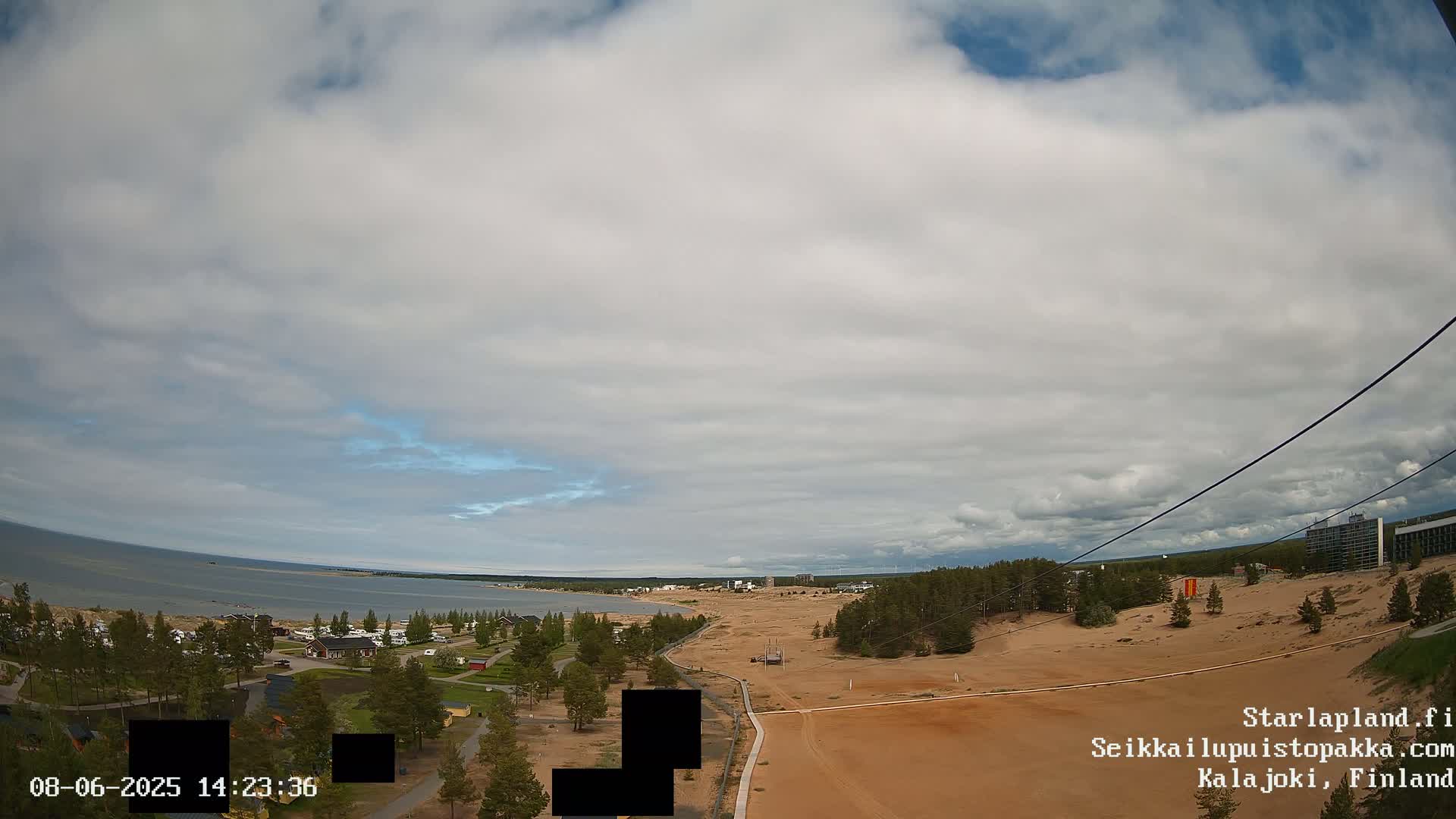 A partly cloudy sky overlooks a sandy landscape with a calm body of water in the distance, featuring sparse vegetation and buildings.