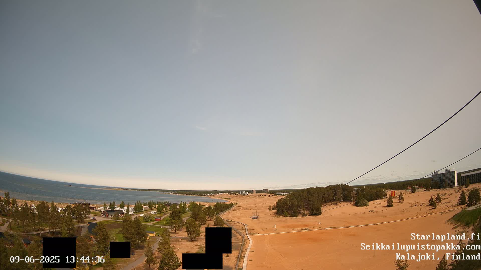 A high-angle view on a clear day shows a sandy landscape near a calm body of water, with some buildings and trees visible in the distance.