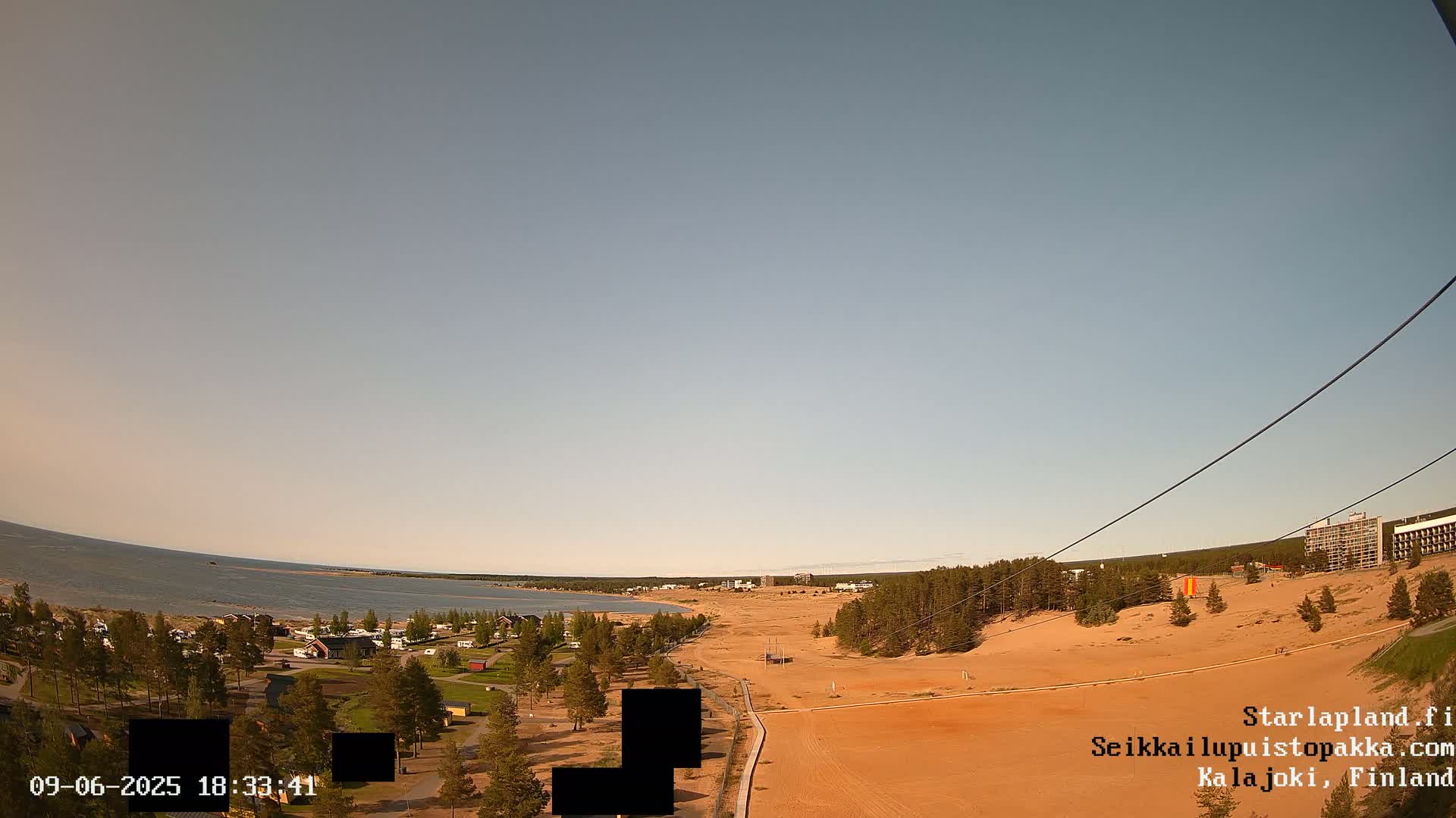 A wide, high-angle view shows a calm body of water, a sandy shoreline with sparse vegetation, and a cluster of buildings under a clear, light-blue sky.