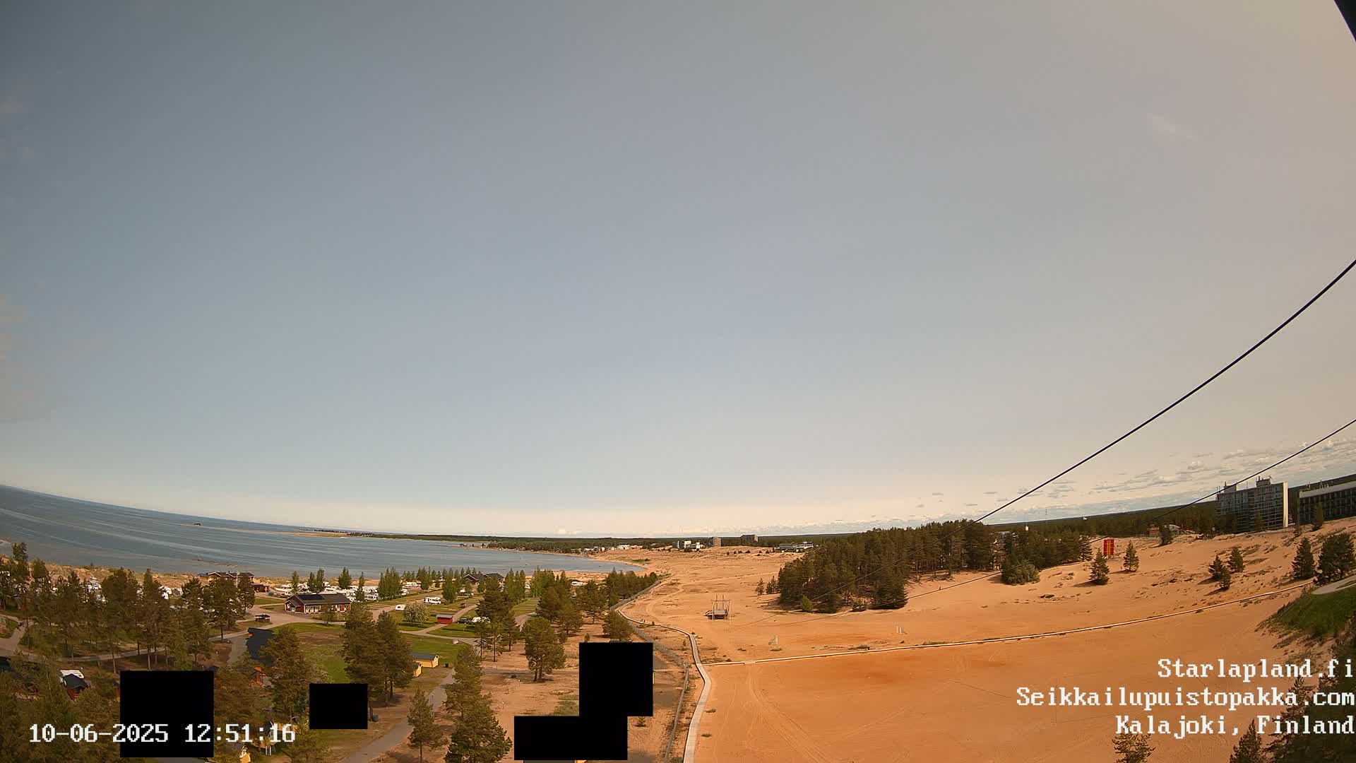 A mostly clear sky overlooks a sandy landscape with a body of water and a sparse collection of buildings in the distance.