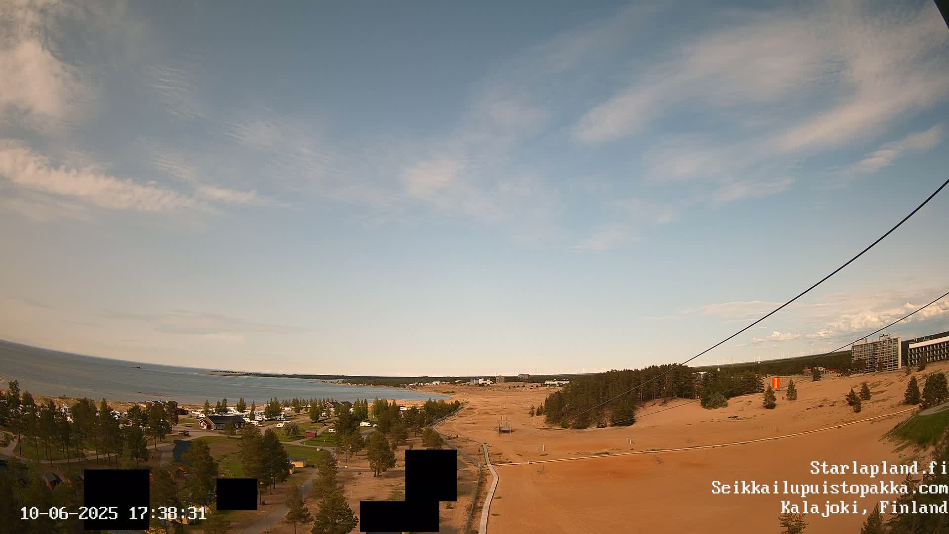 A mostly clear sky overlooks a sandy landscape with sparse vegetation, a body of water in the distance, and a few buildings.