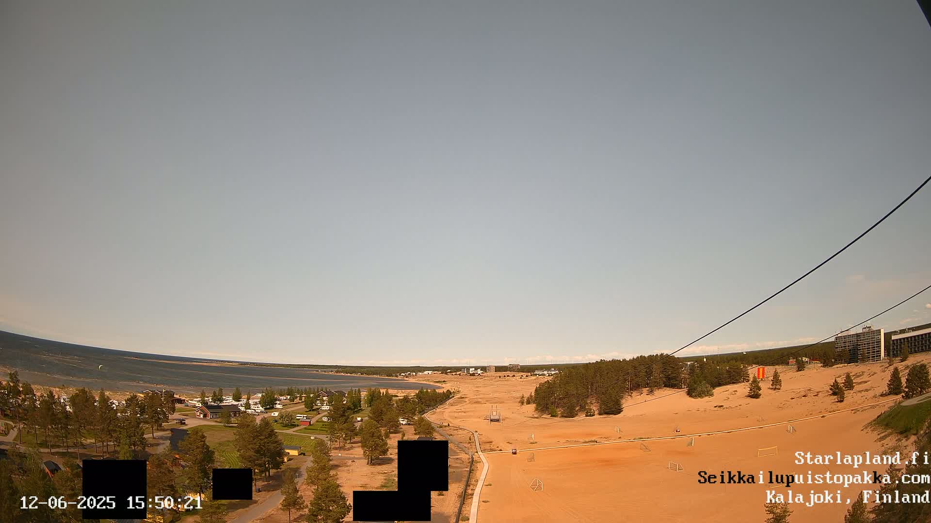 An aerial view shows a sandy area with a small town and a body of water in the background under a clear sky.