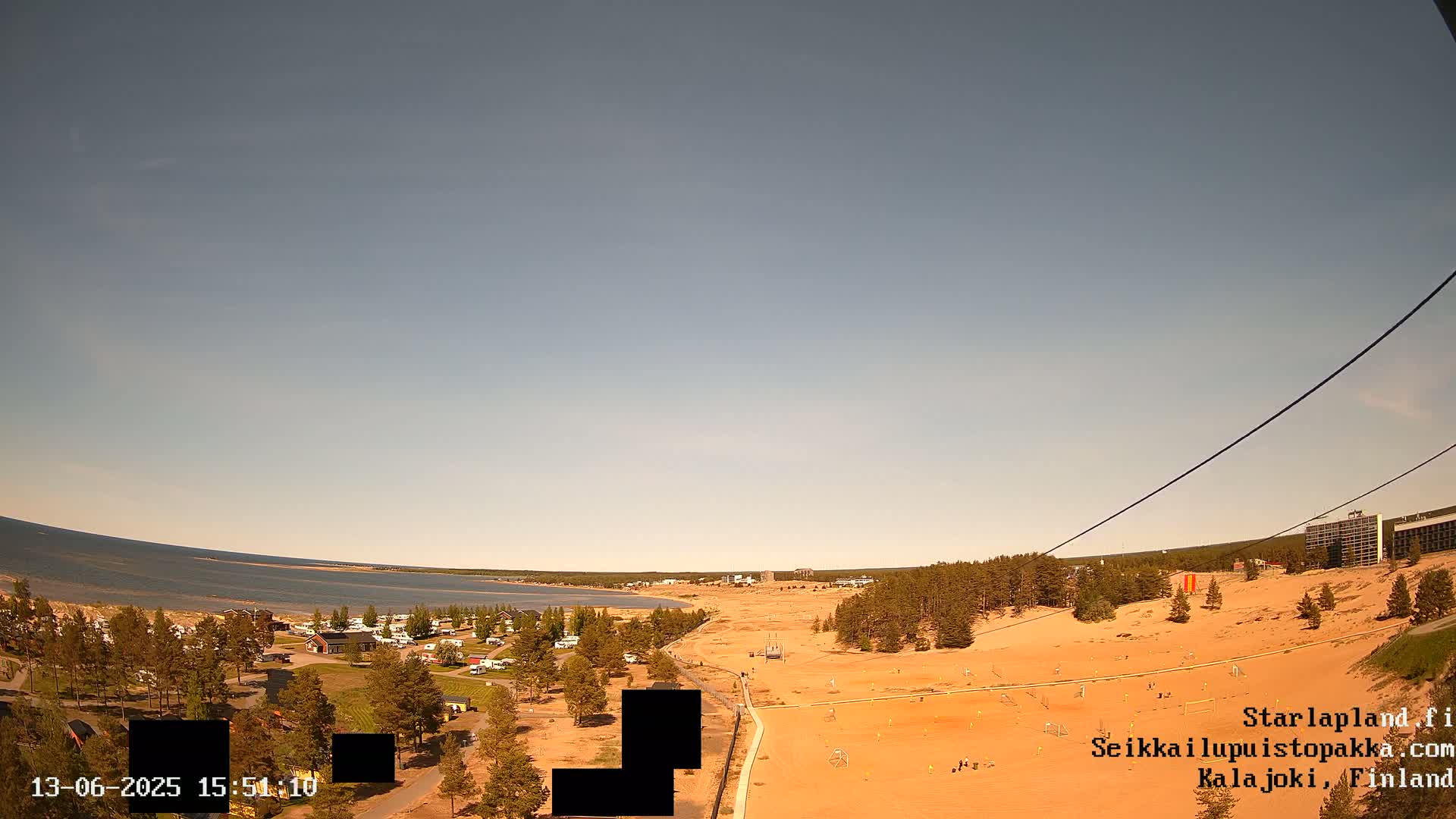 A high-angle, long shot shows a sandy landscape with a calm body of water in the background under a clear, light-blue sky.
