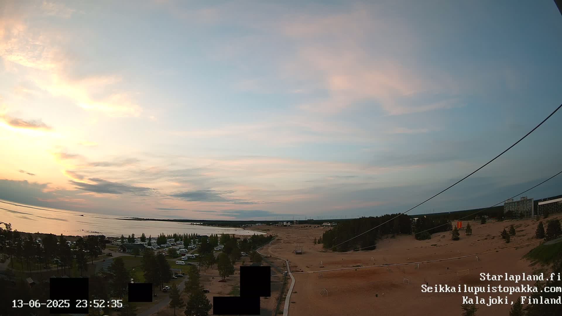 A calm, sunset-lit seaside scene shows a sandy area with a campground and a few buildings in the distance, under a mostly clear sky.