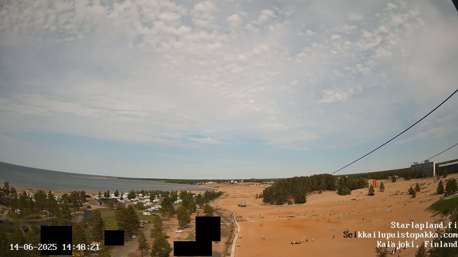 An aerial view shows a sandy area with recreational fields, a campsite, and a calm body of water under a partly cloudy sky.