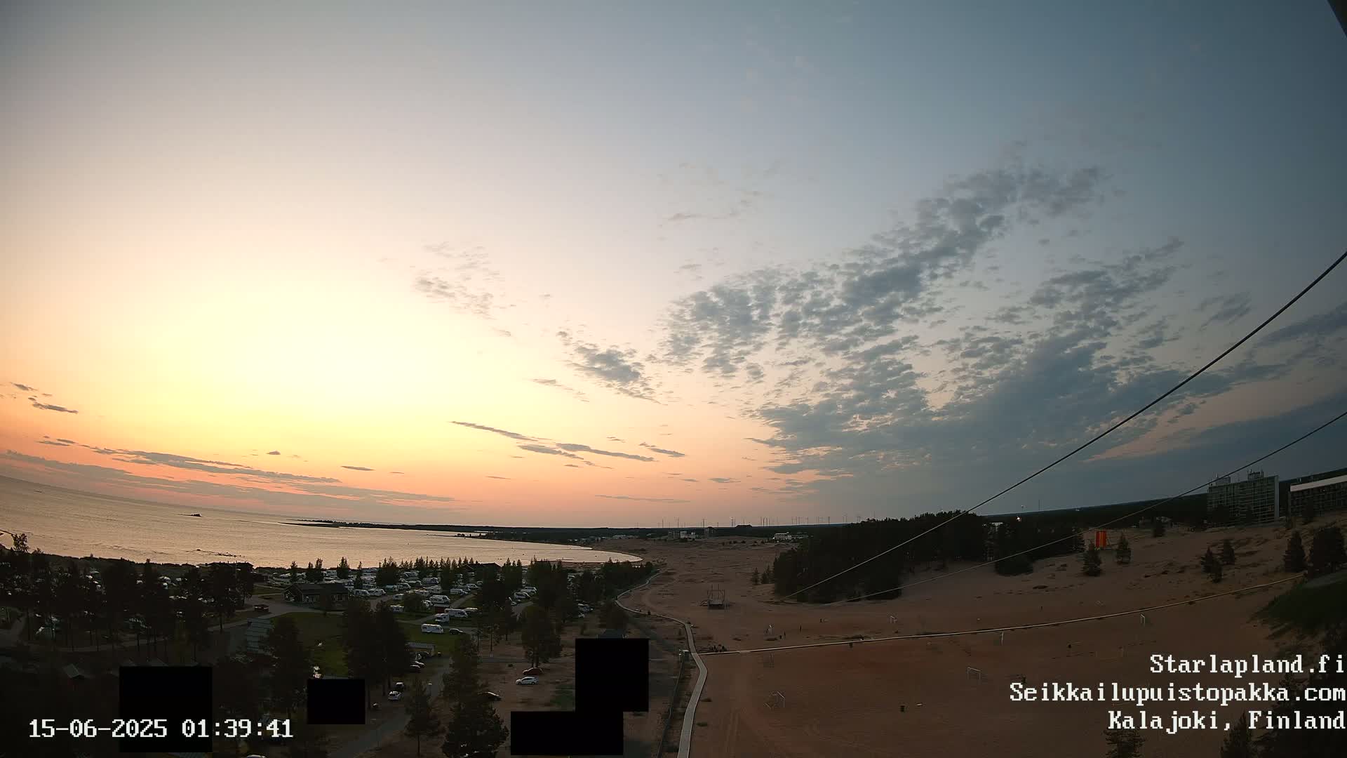 A high-angle view shows a sandy beach, a calm body of water, a campground, and some sparse trees under a mostly cloudy sky.