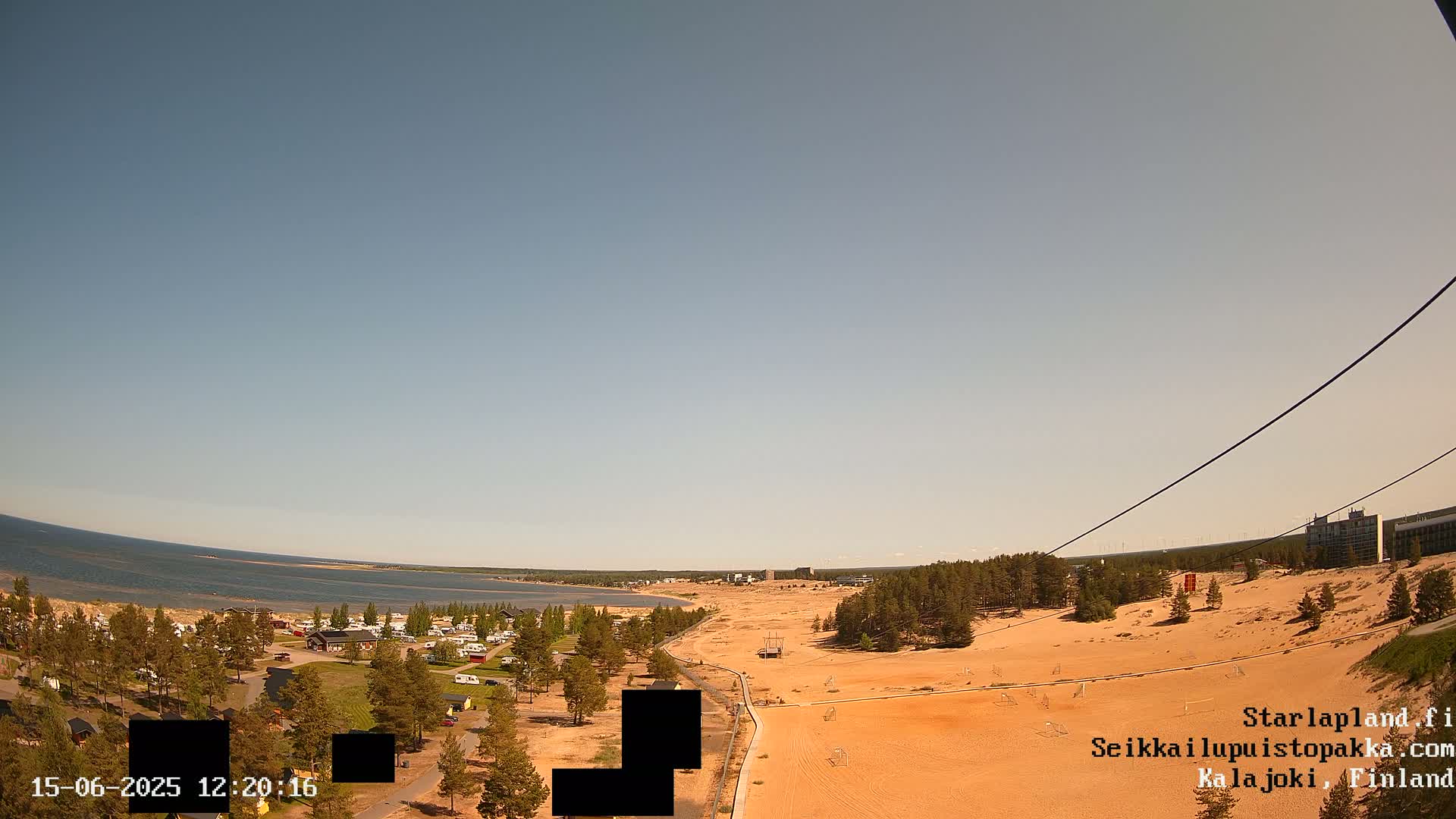 An aerial view shows a sandy landscape with a calm body of water in the distance under a clear blue sky, featuring a campground, sparse trees, and a building in the background.
