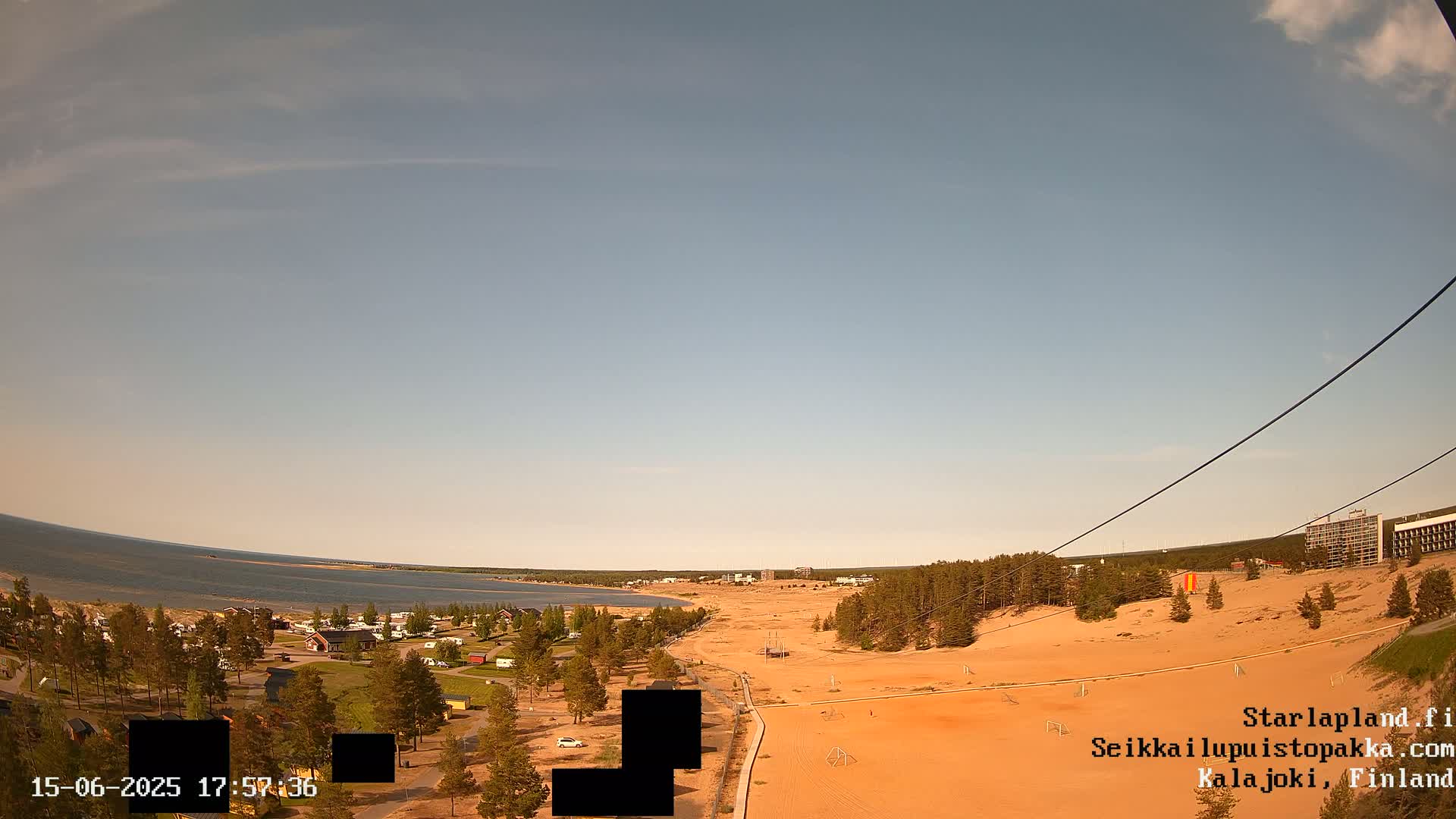 A mostly clear sky overlooks a sandy landscape with a calm body of water in the distance, sparse trees, and buildings visible near the shore.