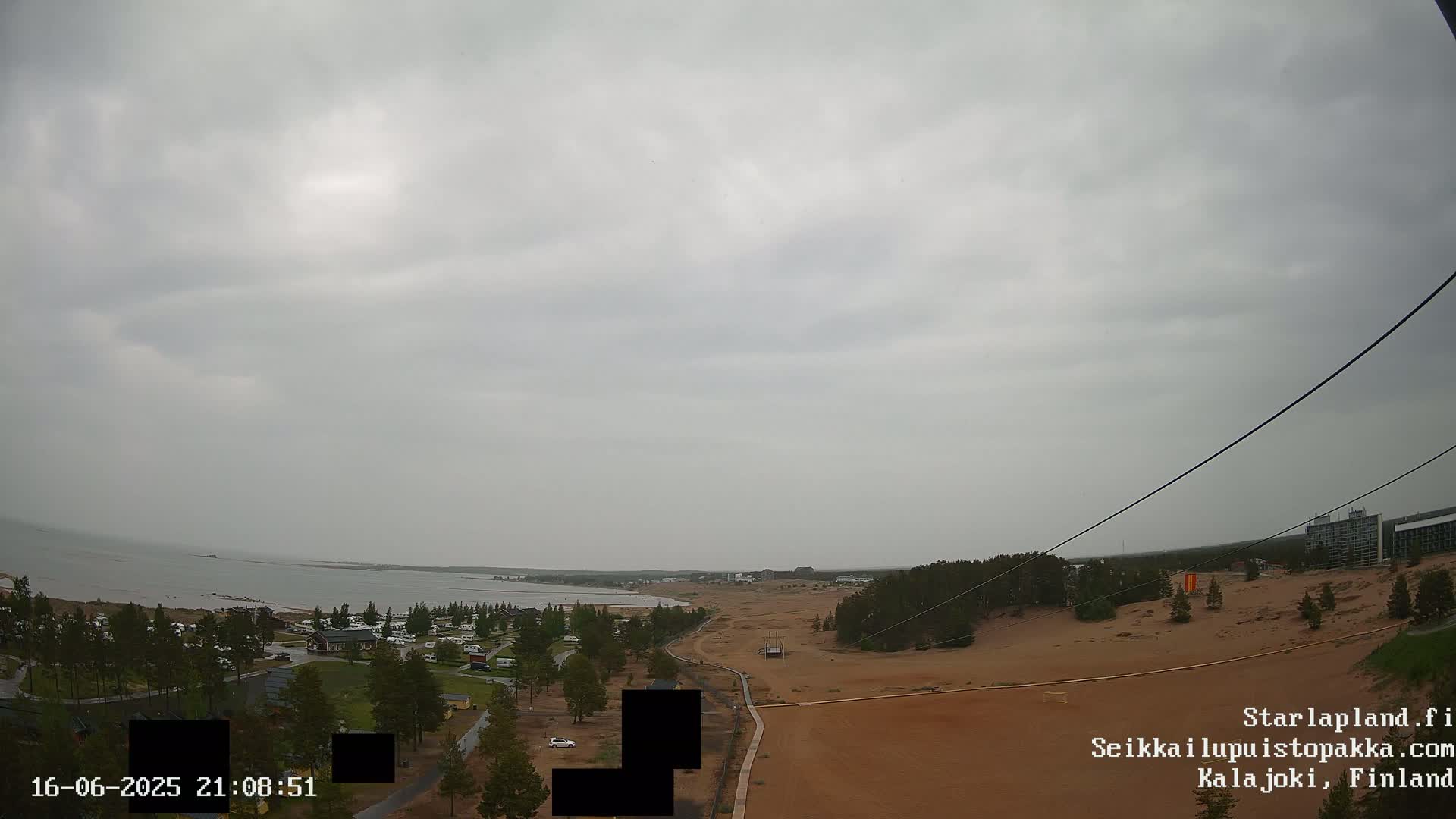 A coastal scene under an overcast sky shows a sandy area with sparse vegetation, a body of water in the distance, and a few buildings visible in the far background.