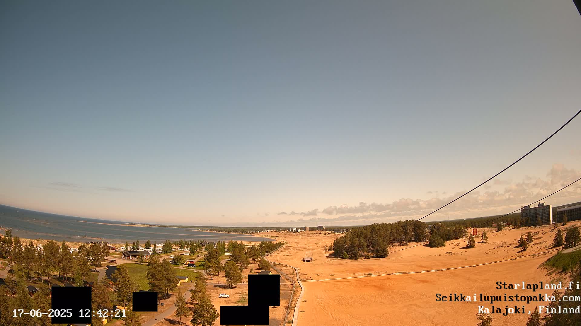 A high-angle, long shot shows a sandy landscape with a body of water and some buildings in the distance under a mostly clear sky.