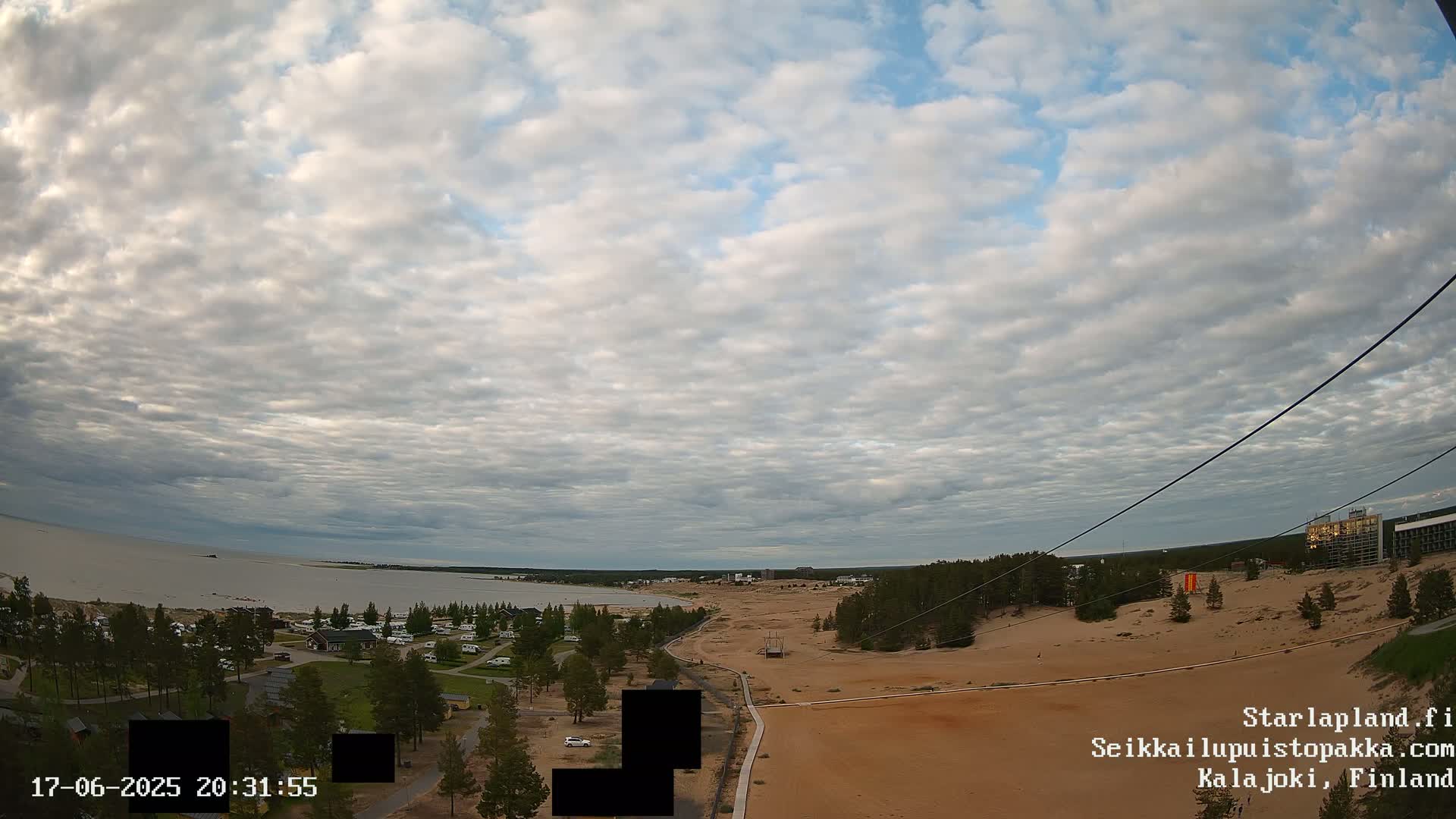 An elevated view shows a calm body of water under a cloudy sky, with a sandy area, vegetation, and buildings in the foreground.