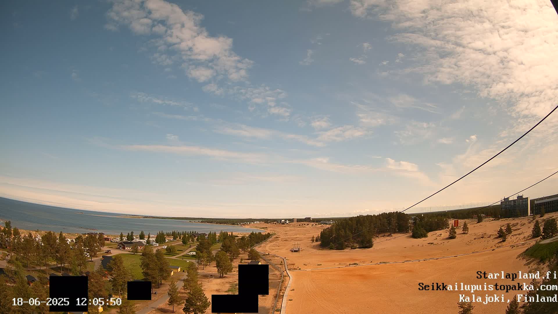 An aerial view shows a sandy landscape with a body of water and a village in the distance under a partly cloudy sky.