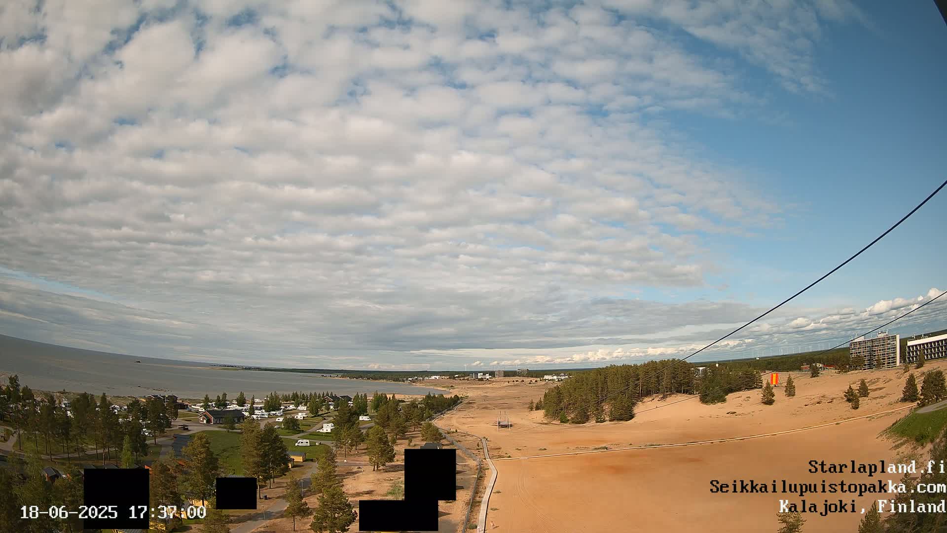A partly cloudy sky overlooks a sandy landscape with a calm body of water and a sparse coastal town in the distance.