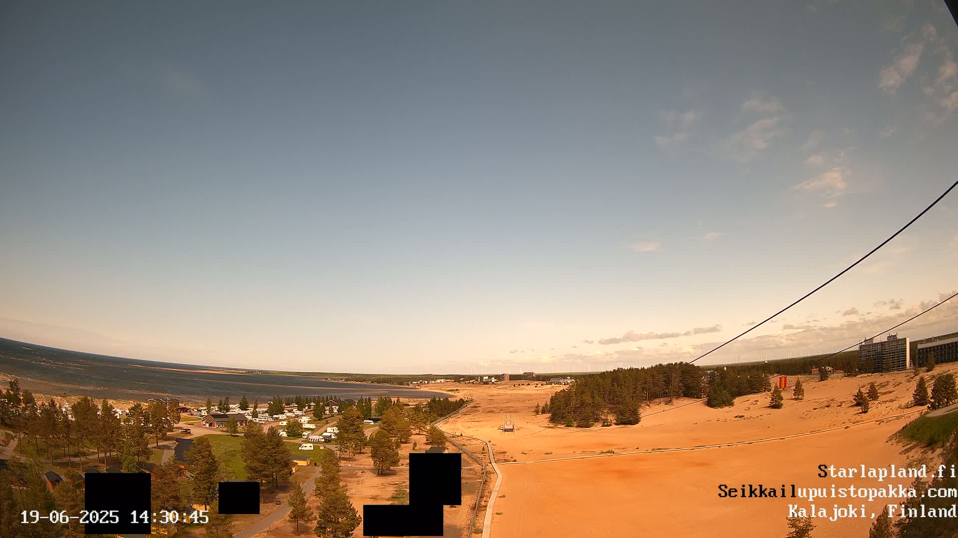 An elevated view shows a sandy landscape with a calm body of water in the background under a mostly clear sky, and a small village with recreational vehicles is visible near the water.