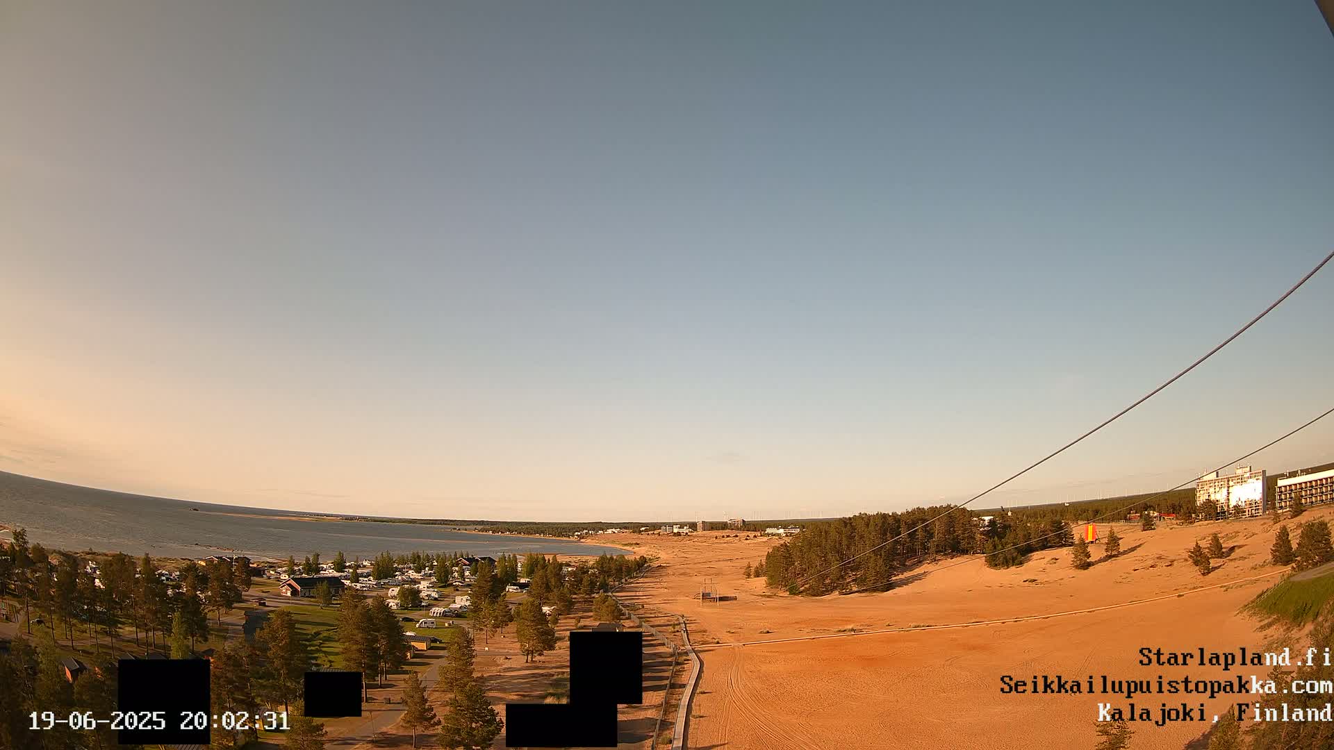 A wide, panoramic view shows a calm body of water, a sandy beach, a campground, and a mostly clear sky under a pale, dusk-like light.