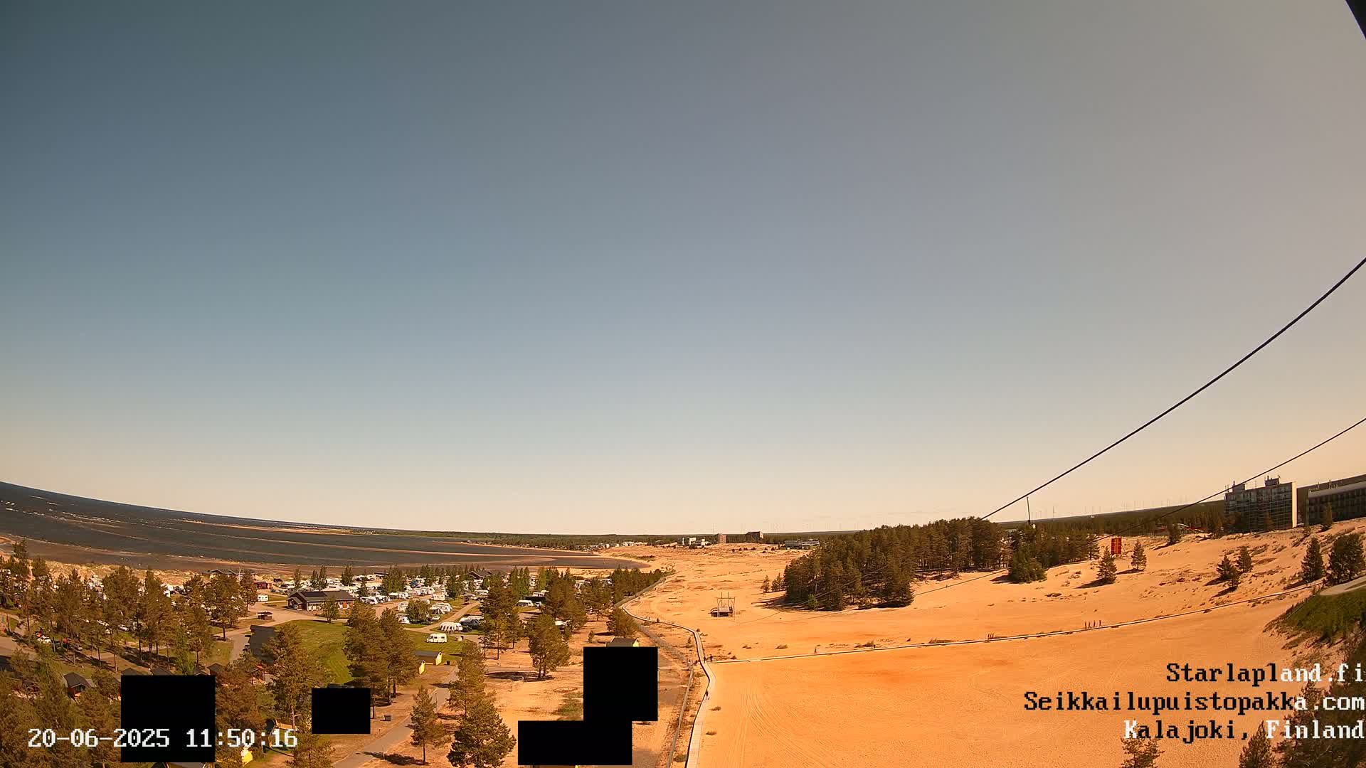 An aerial view shows a sandy landscape with a calm body of water in the background under a clear, sunny sky.