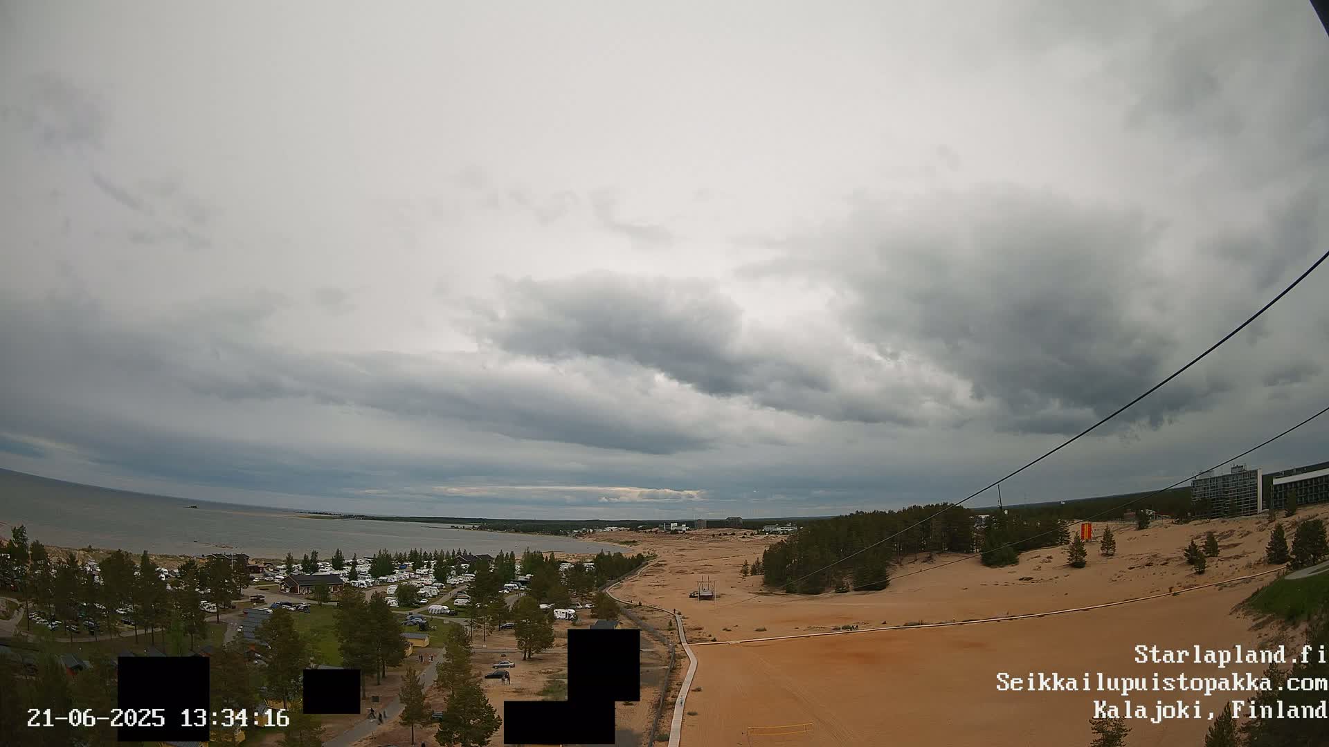 An overcast sky hangs over a sandy landscape next to a calm body of water, with a campsite and buildings visible in the distance.